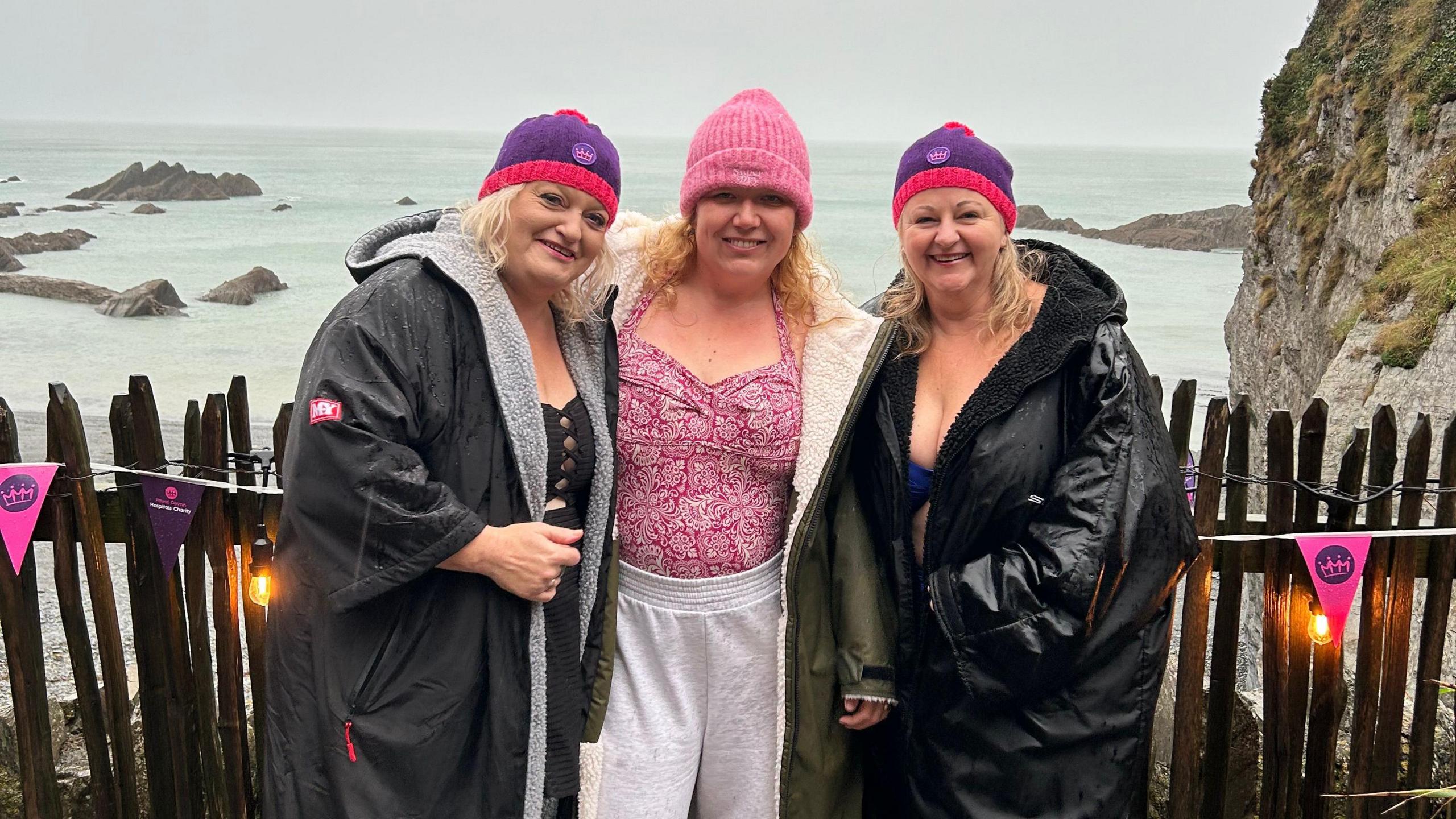 Three women who are all wearing hats and coats, pictured in front of the sea with a number of rocks in the distance.
