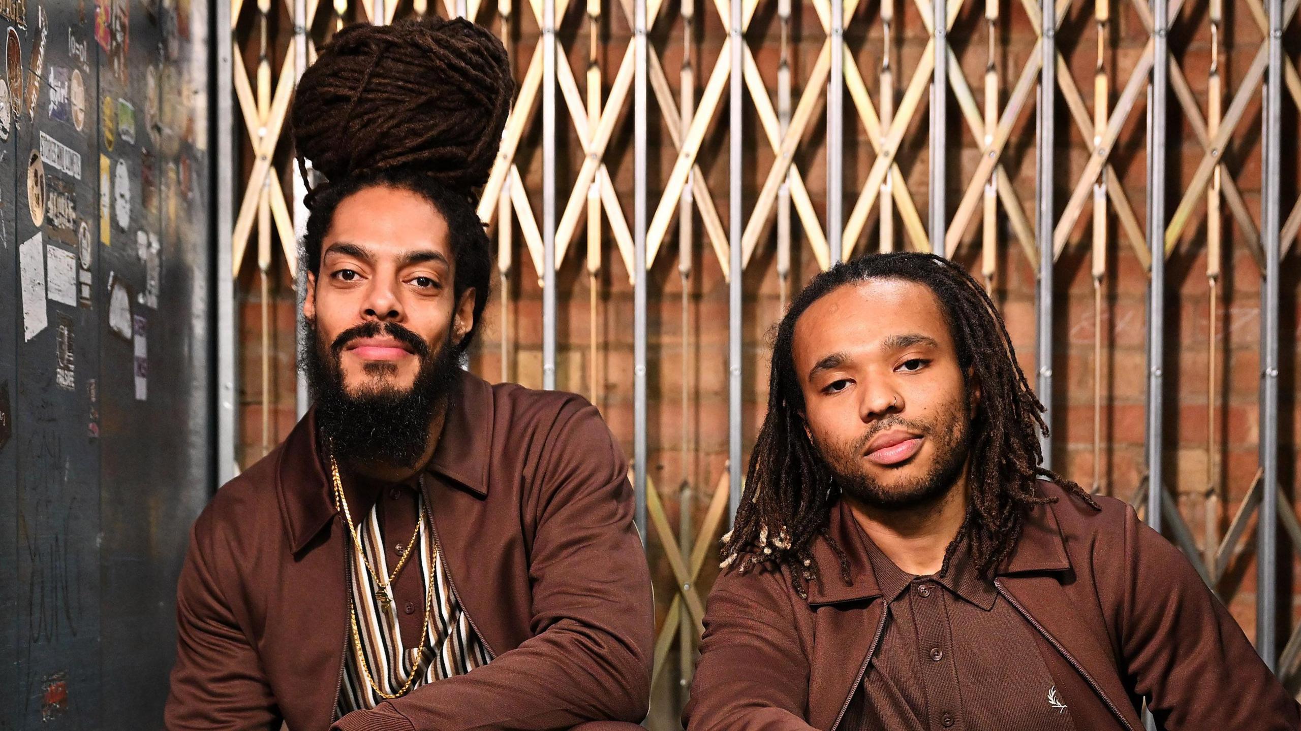 Two men smile at the camera sitting next to each other. One man has dark dreadlocks that sit on top of his head as well as a dark beard. He is wearing a burgandy jacket with a burgandy and white top on underneath. The other man has shoulder length braids and is also wearing a burgandy jacket and matching top underneath.