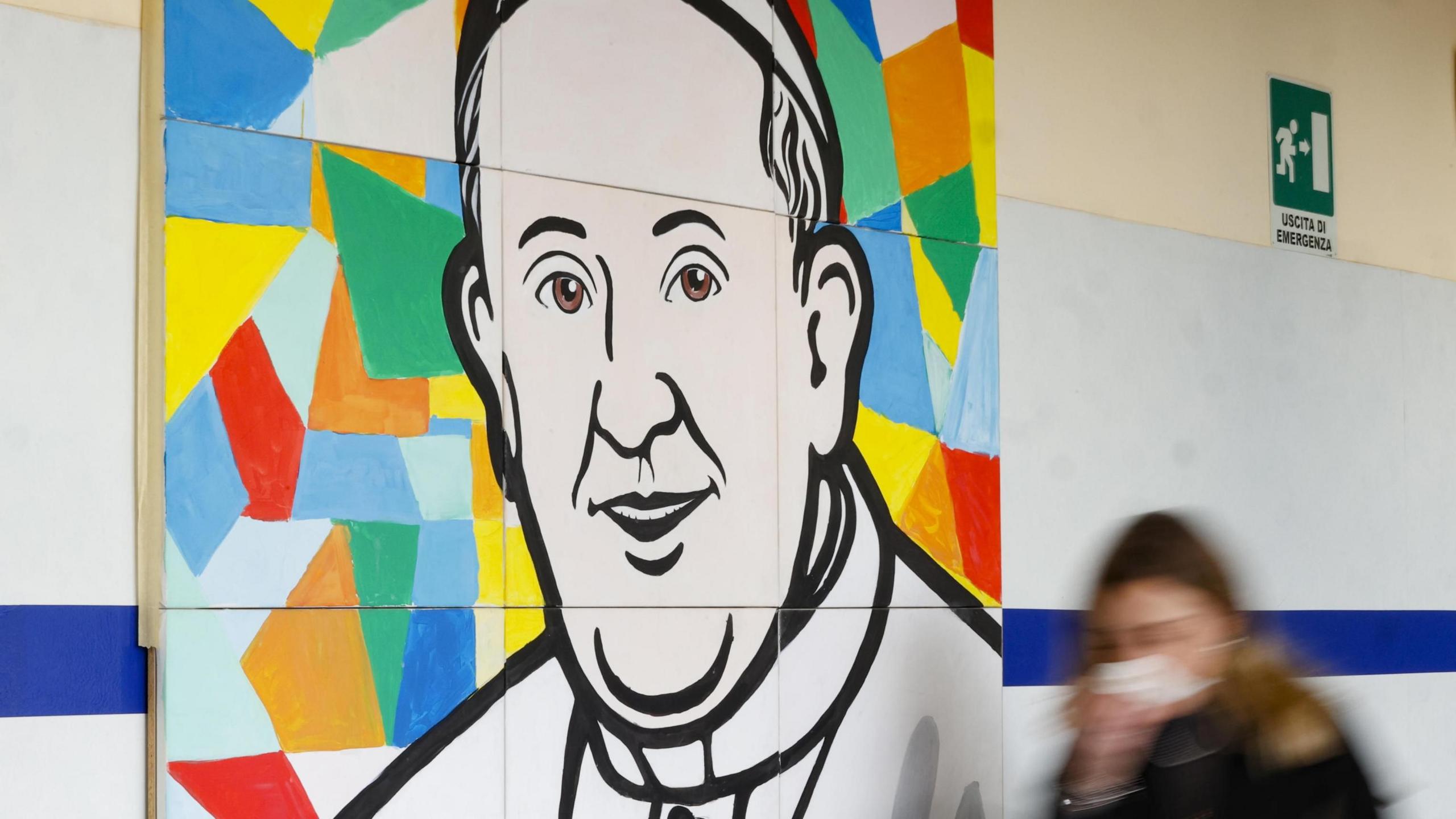 The blurred figure of a woman dressed in black walks past a mural of Pope Francis, which consists of a line drawing of his head and shoulders set against a background of multi-coloured shapes.