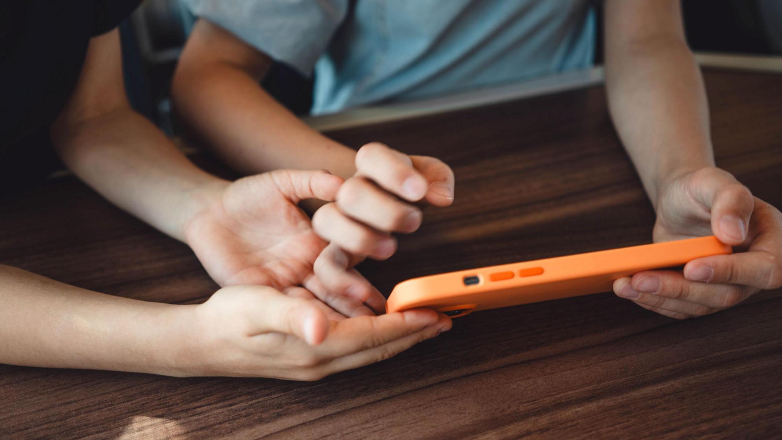 Two children, their faces not visible, hold a smart phone in an orange case on a brown table top.