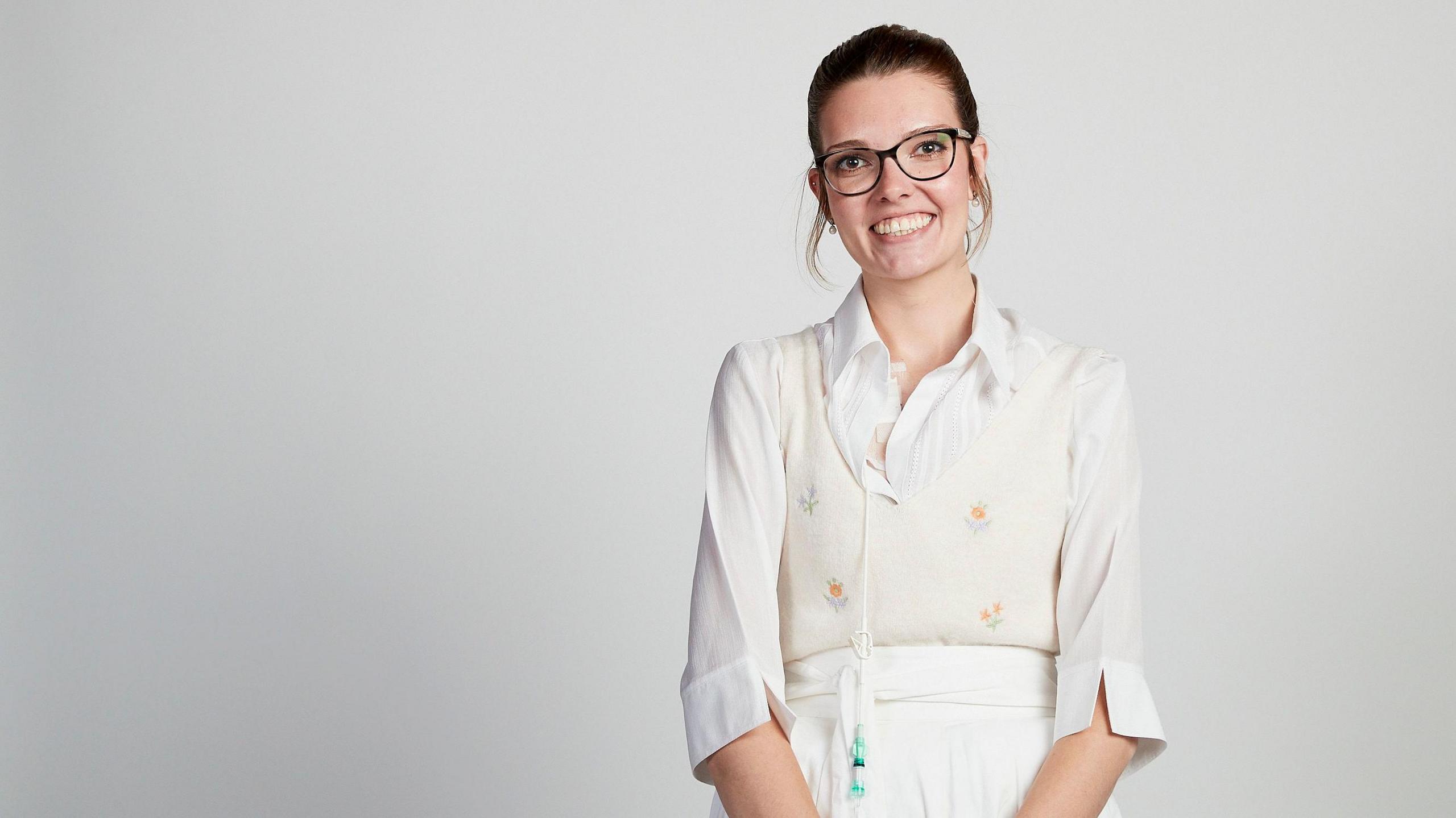 Celia Chartres-Aris smiling for the camera in a white room. She is wearing a dress with flowers. There is a feeding tube hanging on one side.