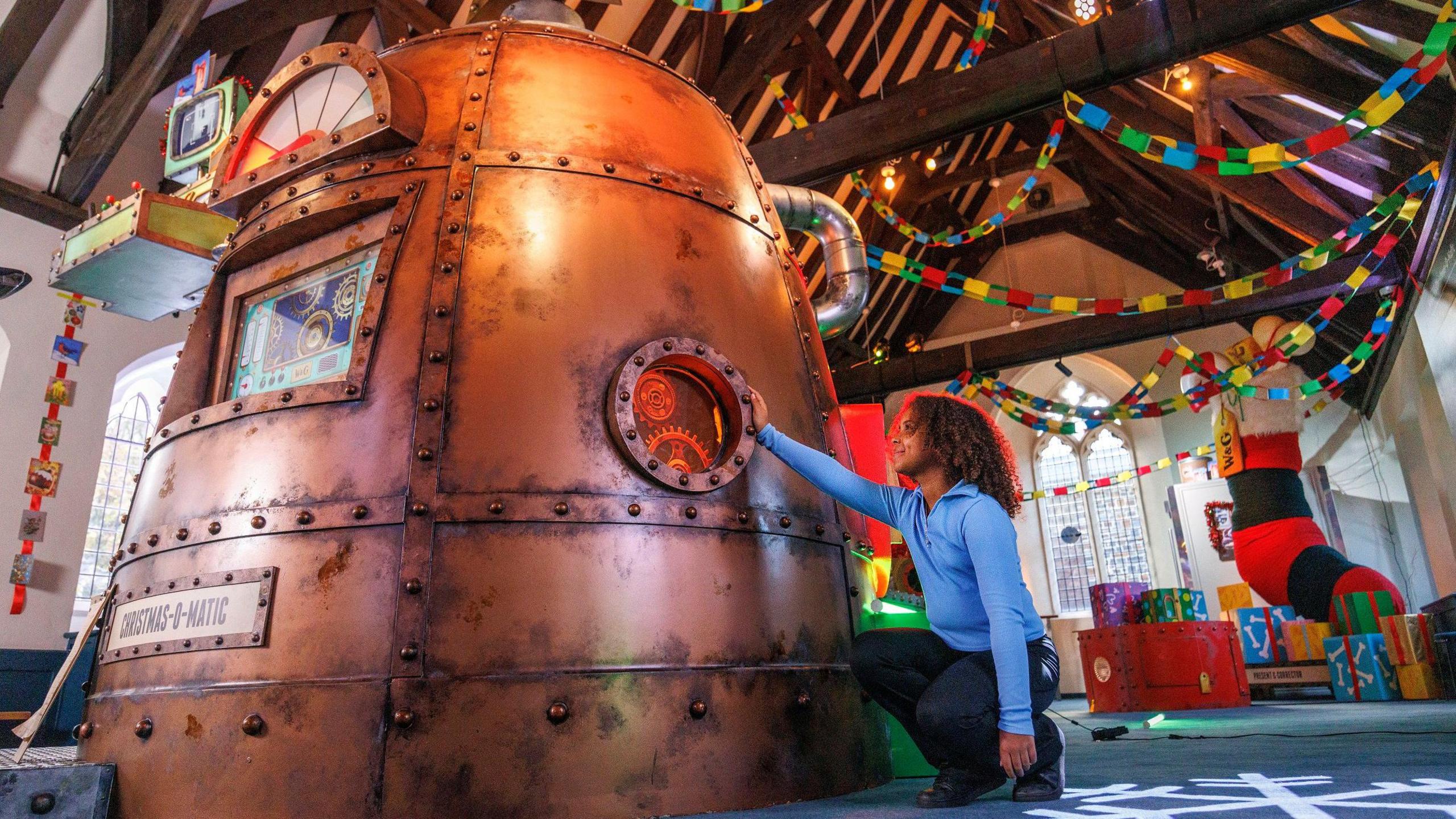 A young child wearing dark trousers and a light blue jumper touches the Christmas-o-Matic, a giant copper dome based on Wallace and Gromit's workshop by Aardman Animations. It is inside the Friary at Cabot Circus and behind it festive streamers and toys can be seen