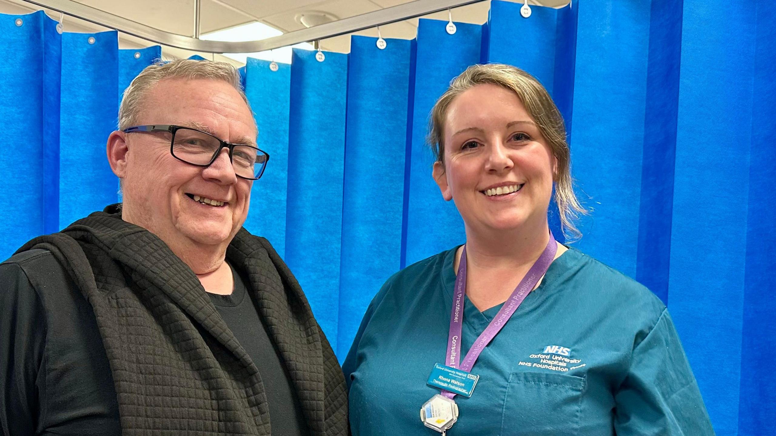 Graham Best is standing with Rhona Watson in front of a blue hospital curtain.