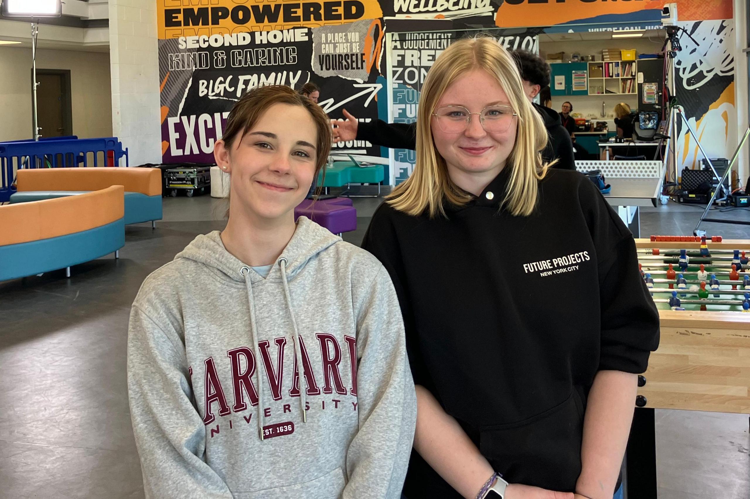 Two girls smiling at the camera as they pose for a picture in a youth club. Maisie on the left has brown hair pulled back in a pony tail and is wearing a grey hoodie with Harvard University branding in large red type. Iga on the right wears a black hoodie bunched up on her arms. She has shoulder-length blond hair and has rimless glasses.