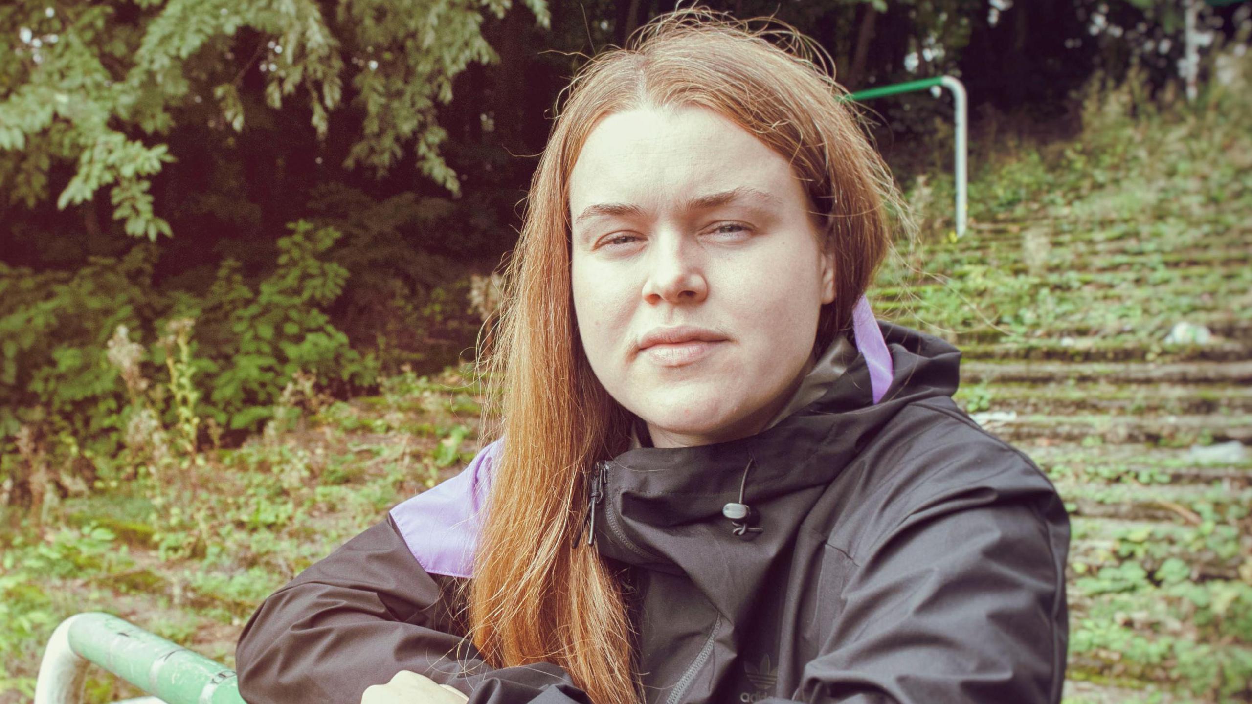 A woman with red hair looks into the camera while standing outside on an old football terrace