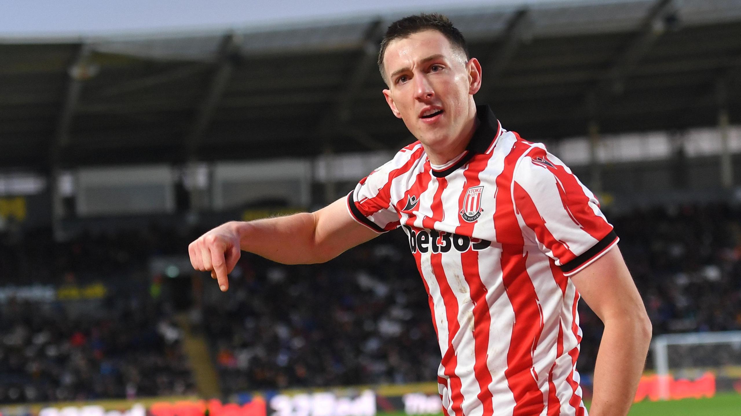 Robert Bozenik looks to his left and holds his right arm out with his index finger pointing to the ground. He is wearing a red and white striped Stoke City kit and running on the pitch during a game after scoring a goal