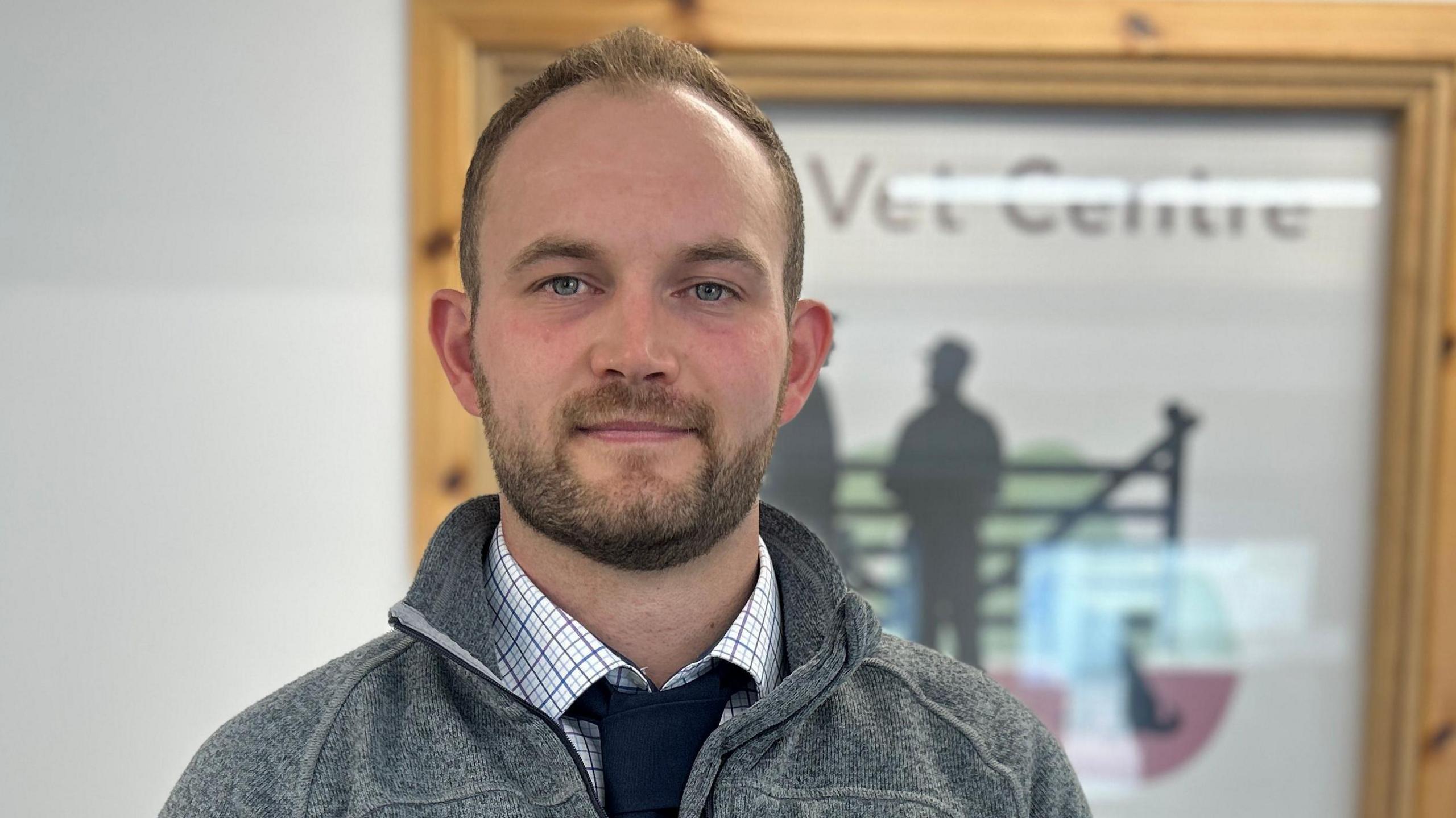 A young man with thin fair hair and a beard, wearing a fleece over a checked shirt and a black tie. He is staring into the camera and is standing in front of a sign that says "vet centre".