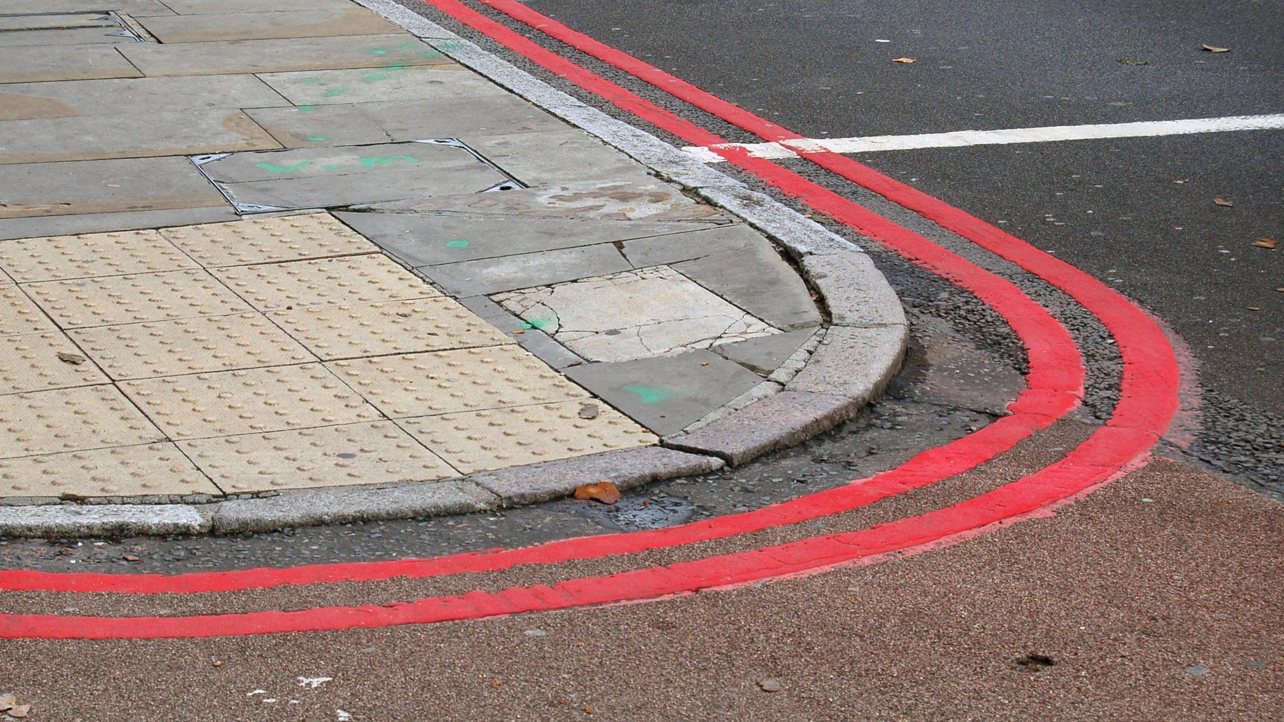 Double red lines curve around a pavement, with tactile paving in the lower part alongside a dropped kerb.