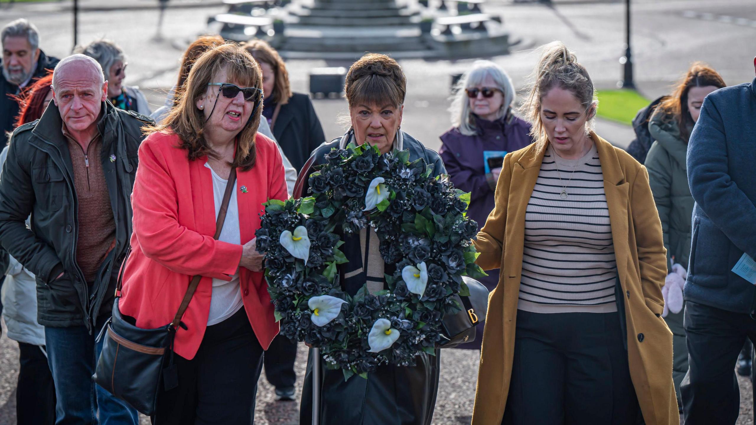 Maria Lynskey holding a black wreath with five white lilies, supported by Dympna Kerr and a sister of 