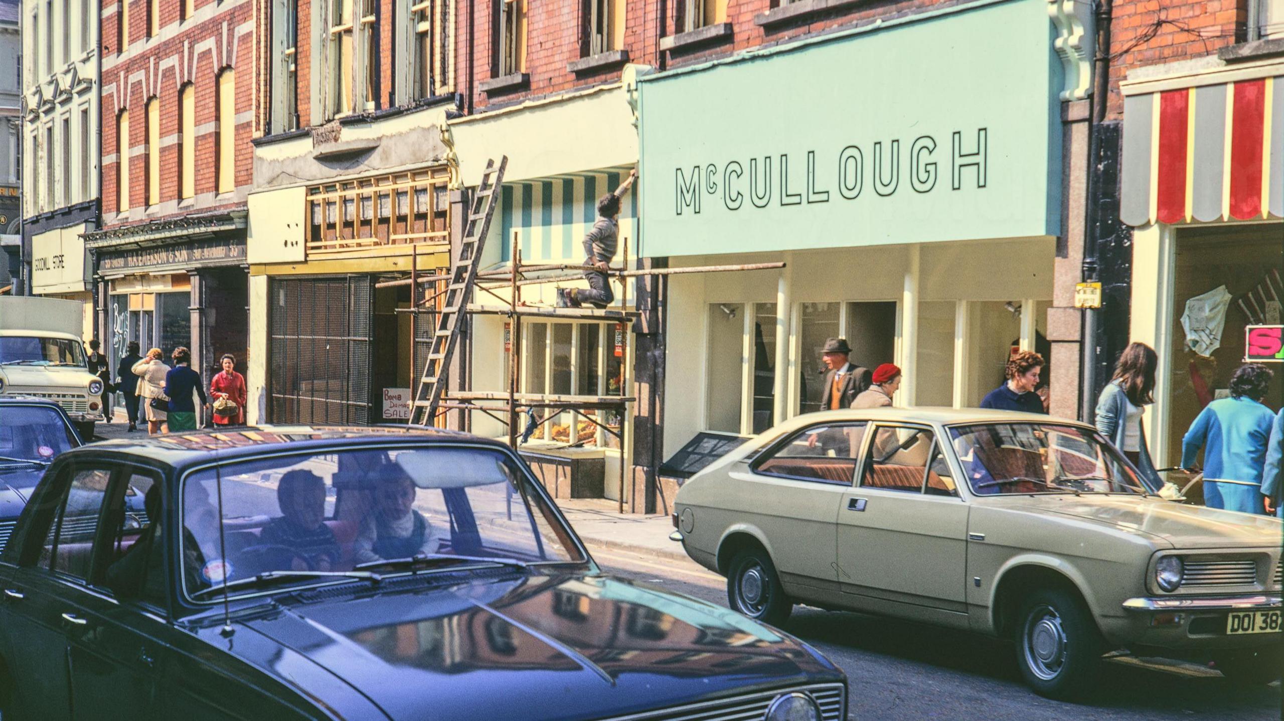 A digitalised photo from a film of a busy street. Shop fronts are being built on scaffolding. Cars are parked and driving along the street. People can be seen looking in shop windows. A view looking west at the top of Scotch Street showing bomb damaged shops with boarded up windows and signs indicating 'business as usual' and bomb damage sale. Shoppers walk the footpath while a man on scaffolding works on a shop front, Shops include 'Goodwill Shop, Emerson and McCullough.