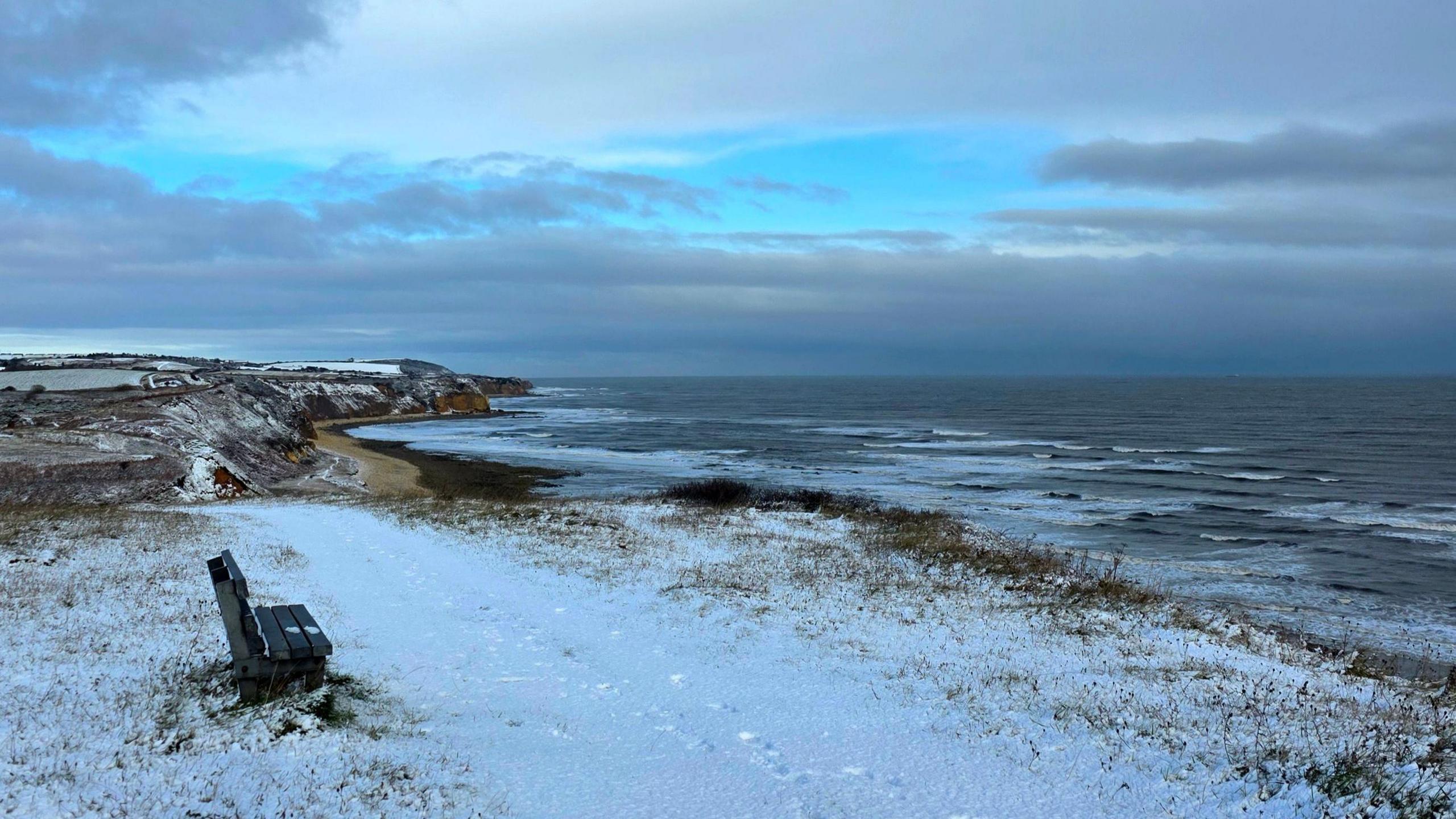 A bench at the top of snow-covered cliffs overlooks the sea waves and the dark blue sky. A beach is visible in the distance.