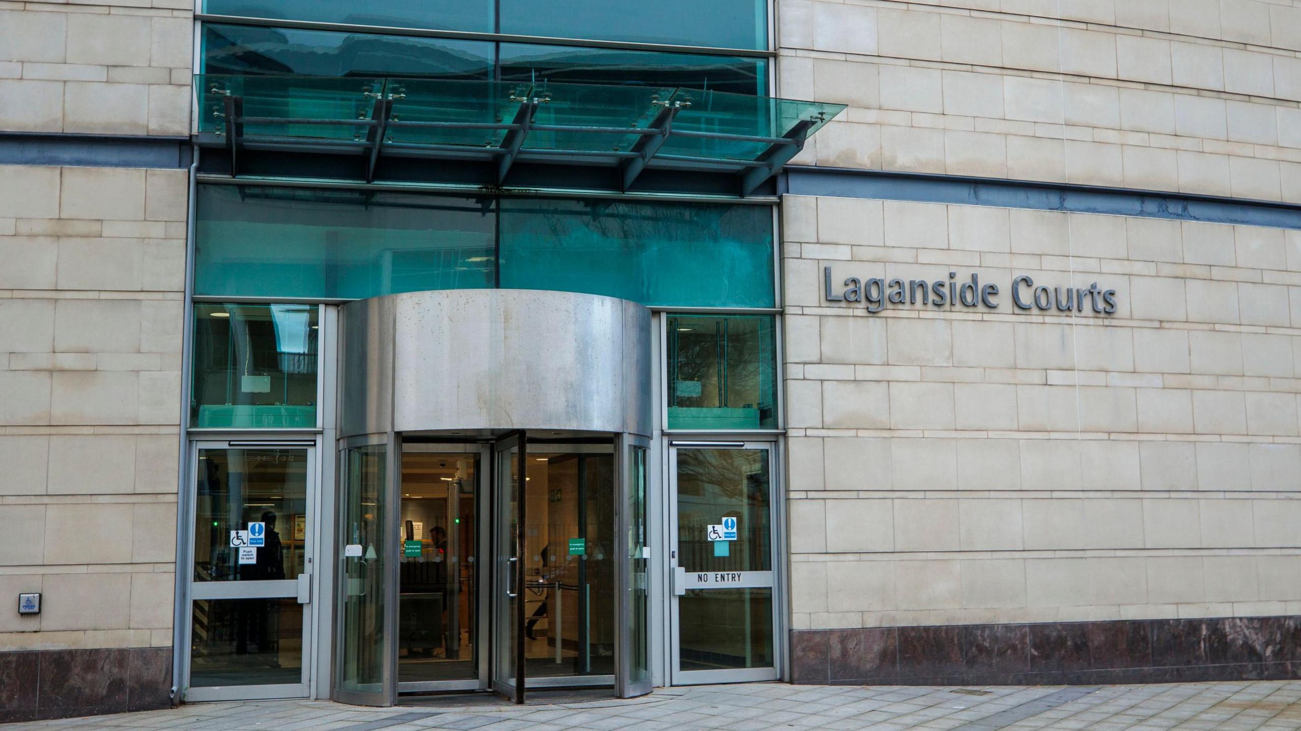A sandstone building with a silver and glass revolving door, and blue tinted glass above it. On the wall next to the door is 'Laganside Courts' written in dark grey writing. The pavement is a light grey stone.
