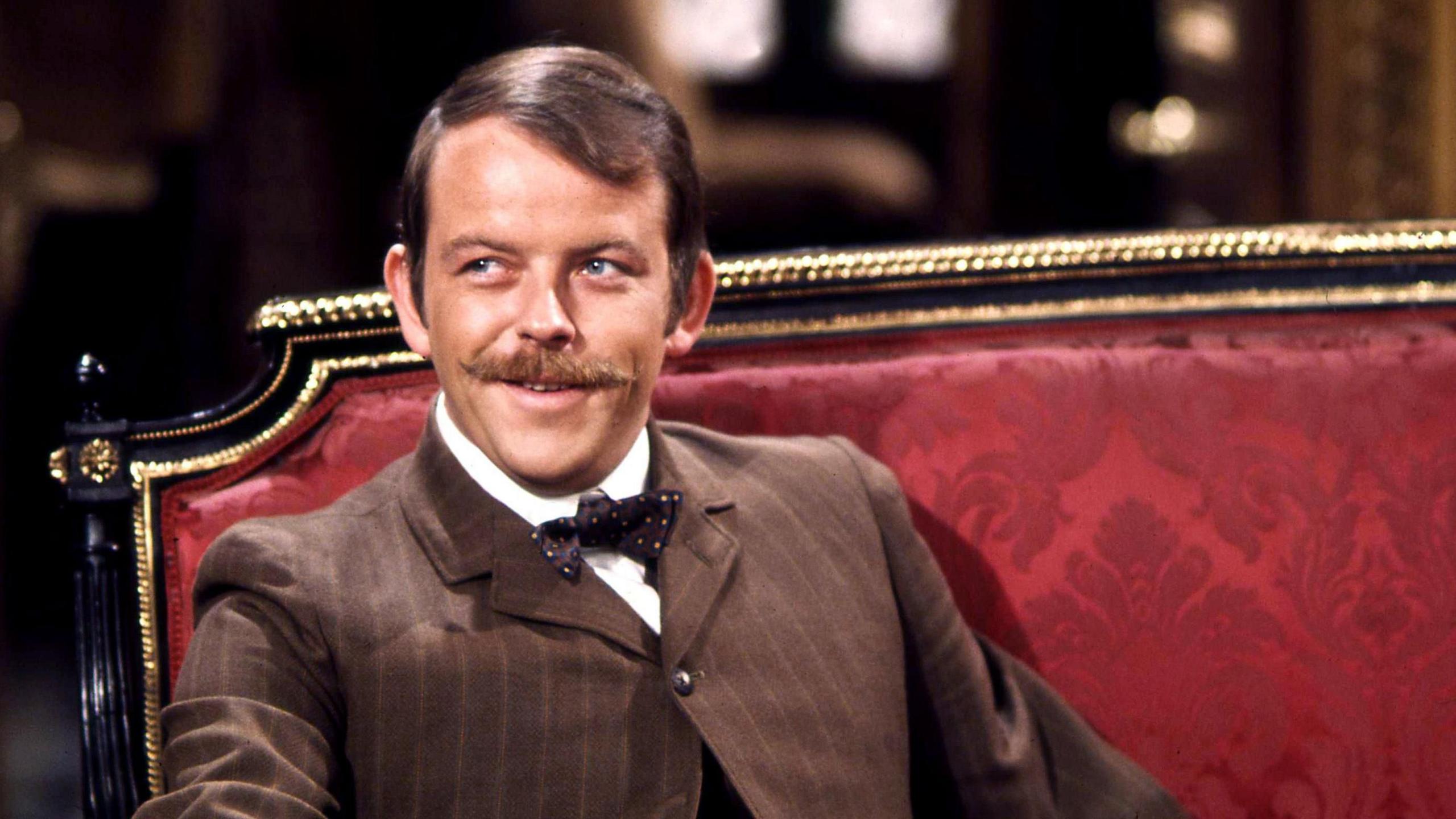 Julian Holloway in character as Algernon Moncrieff. He has a large moustache and is wearing a brown pinstripe suit with a bow tie. He is sitting on a black and gold chair upholstered with crimson brocade.