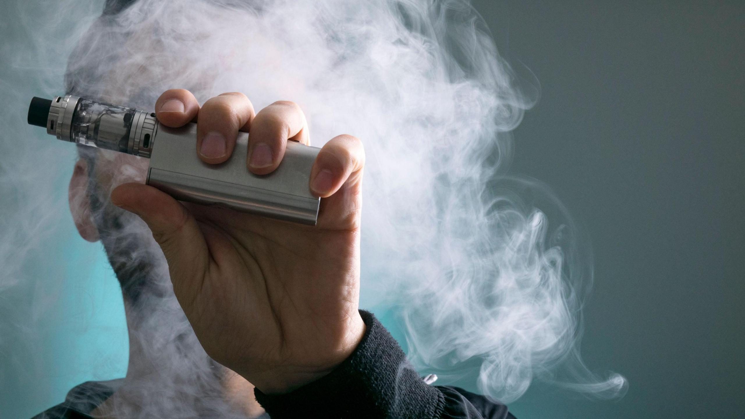 A silver vaping device being held close to the camera. A man's face is obscured by the device and a cloud of vapour.