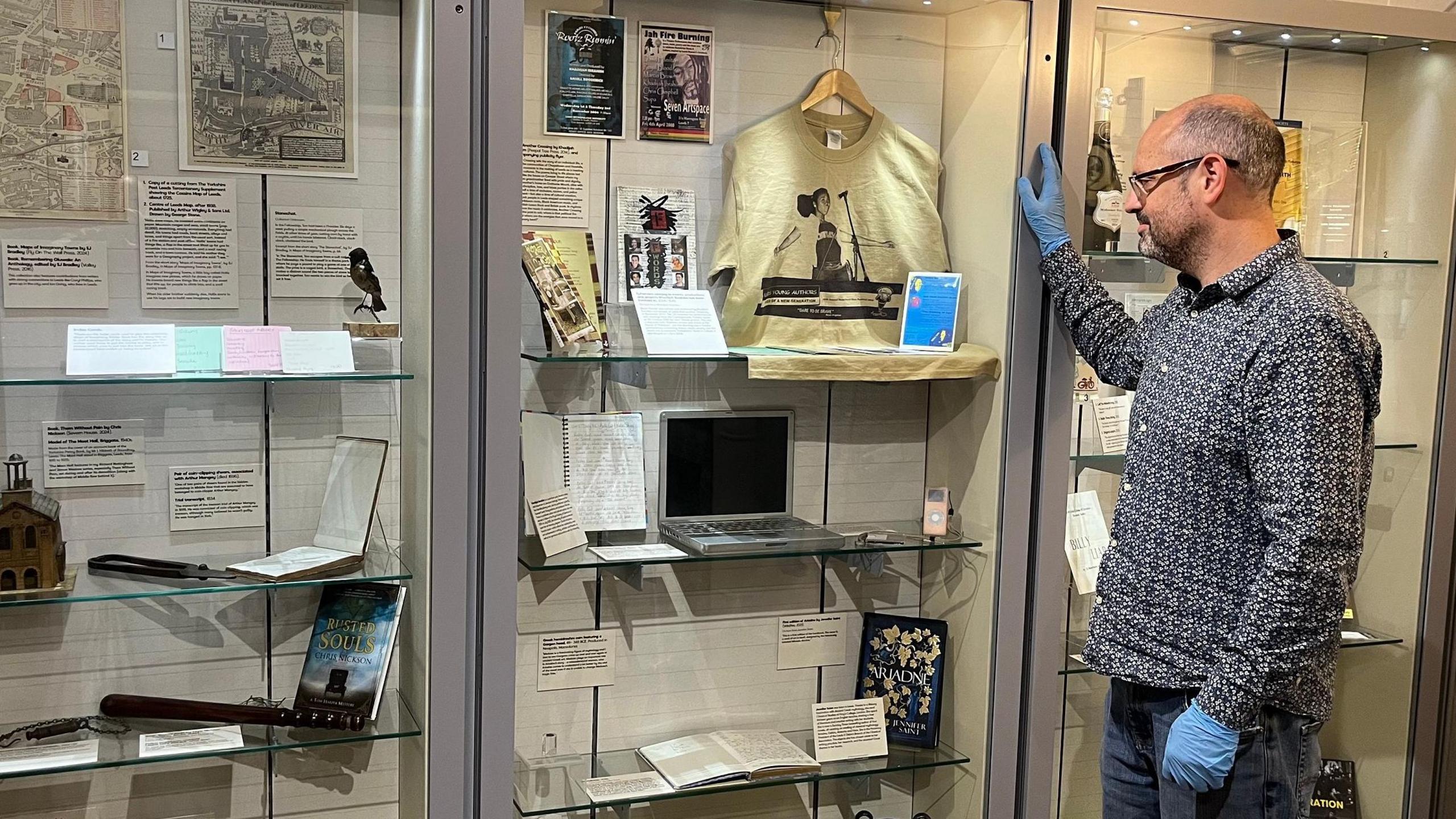 A man with a blue shirt and gloves peers into a glass cabinet in a museum