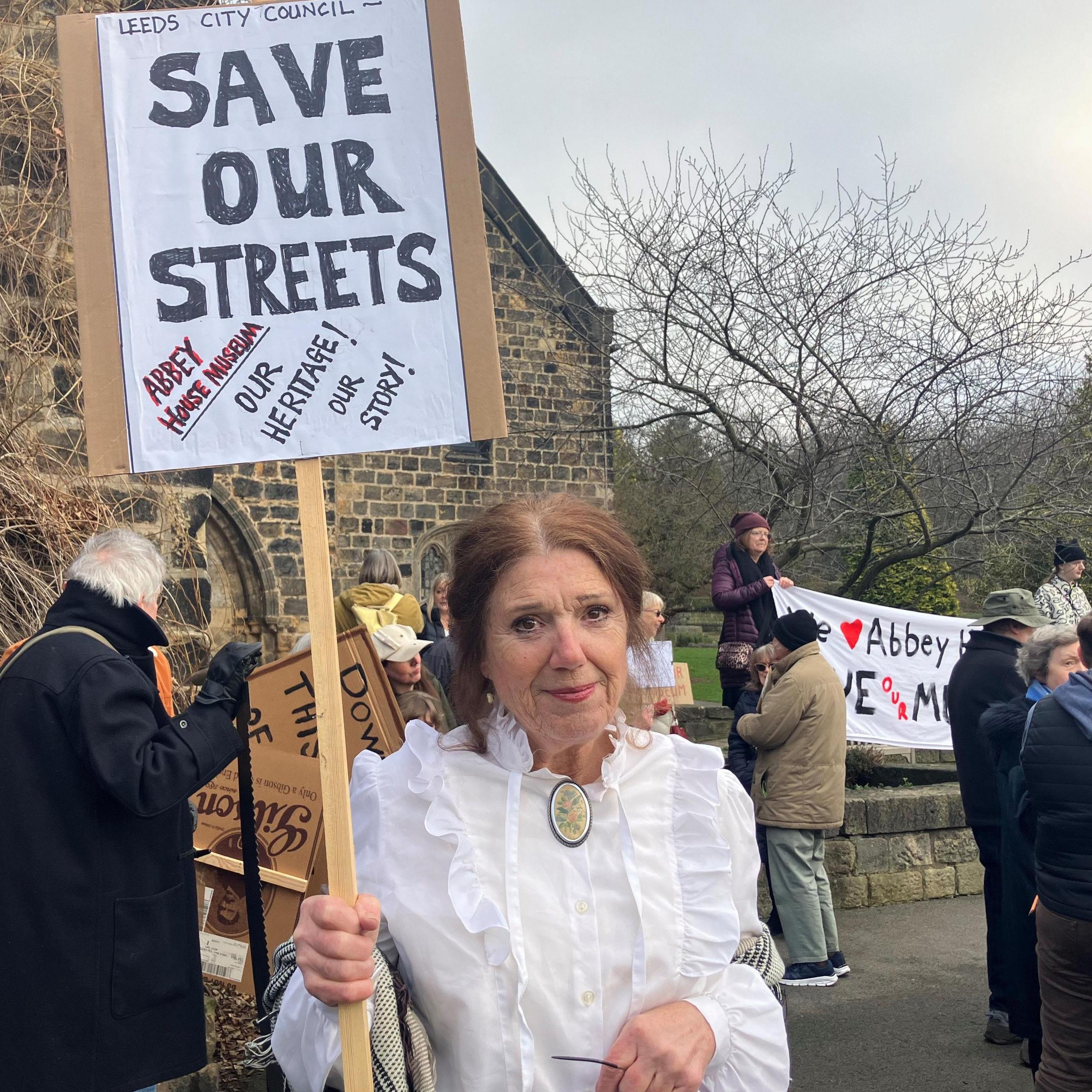 A woman wearing Victorian costume - a white blouse with frills and a neck clasp - holds a placard reading "Save our Streets". She is standing with fellow protesters in front of a museum building.