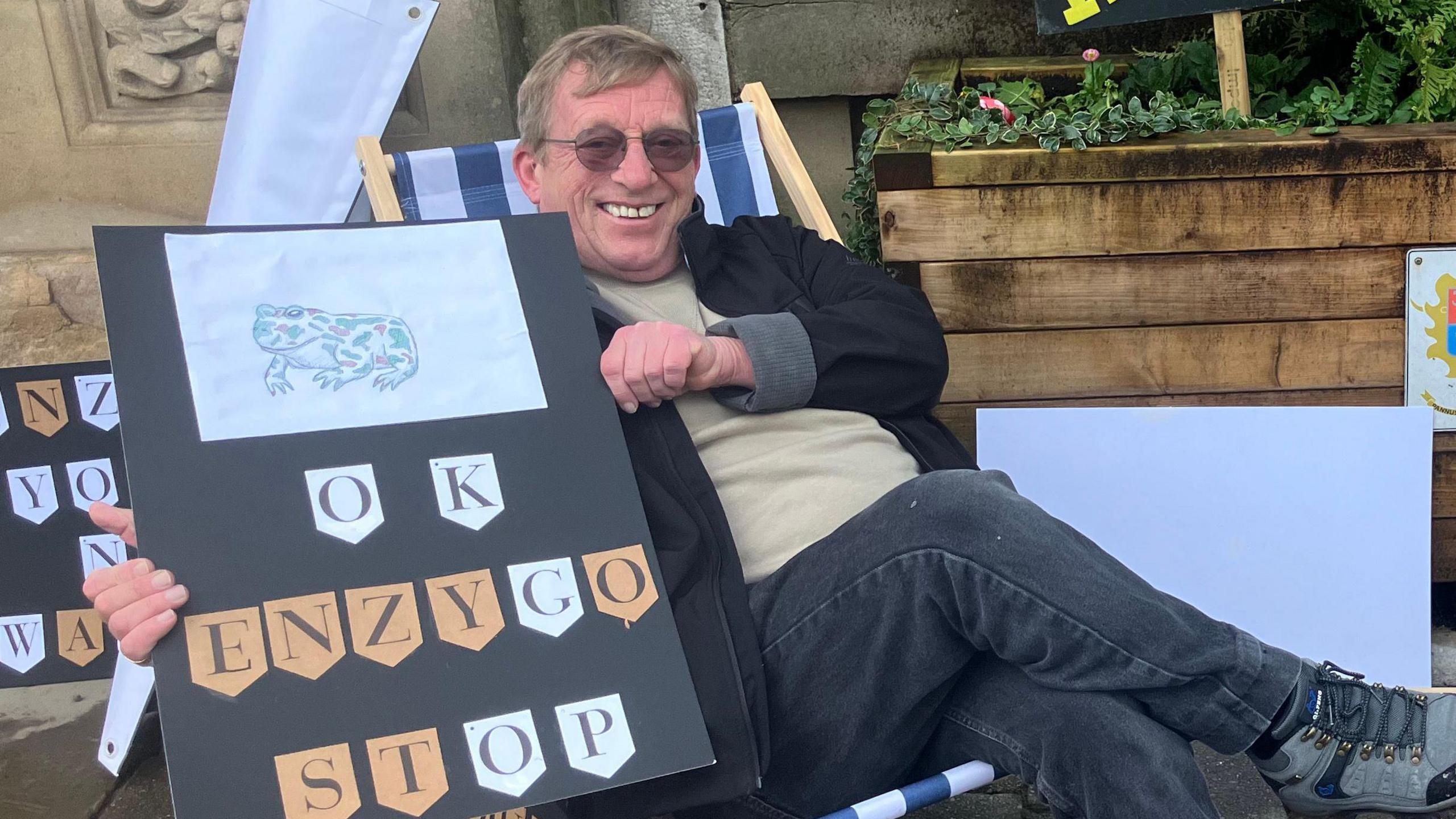 Ged Andrews with grey hair, glasses, wearing a black jacket, beige top and black jeans, sits in a deckchair smiling. He is holding up a black post with a picture of a frog on and wording underneath reading 'OK Enzygo Stop'. 