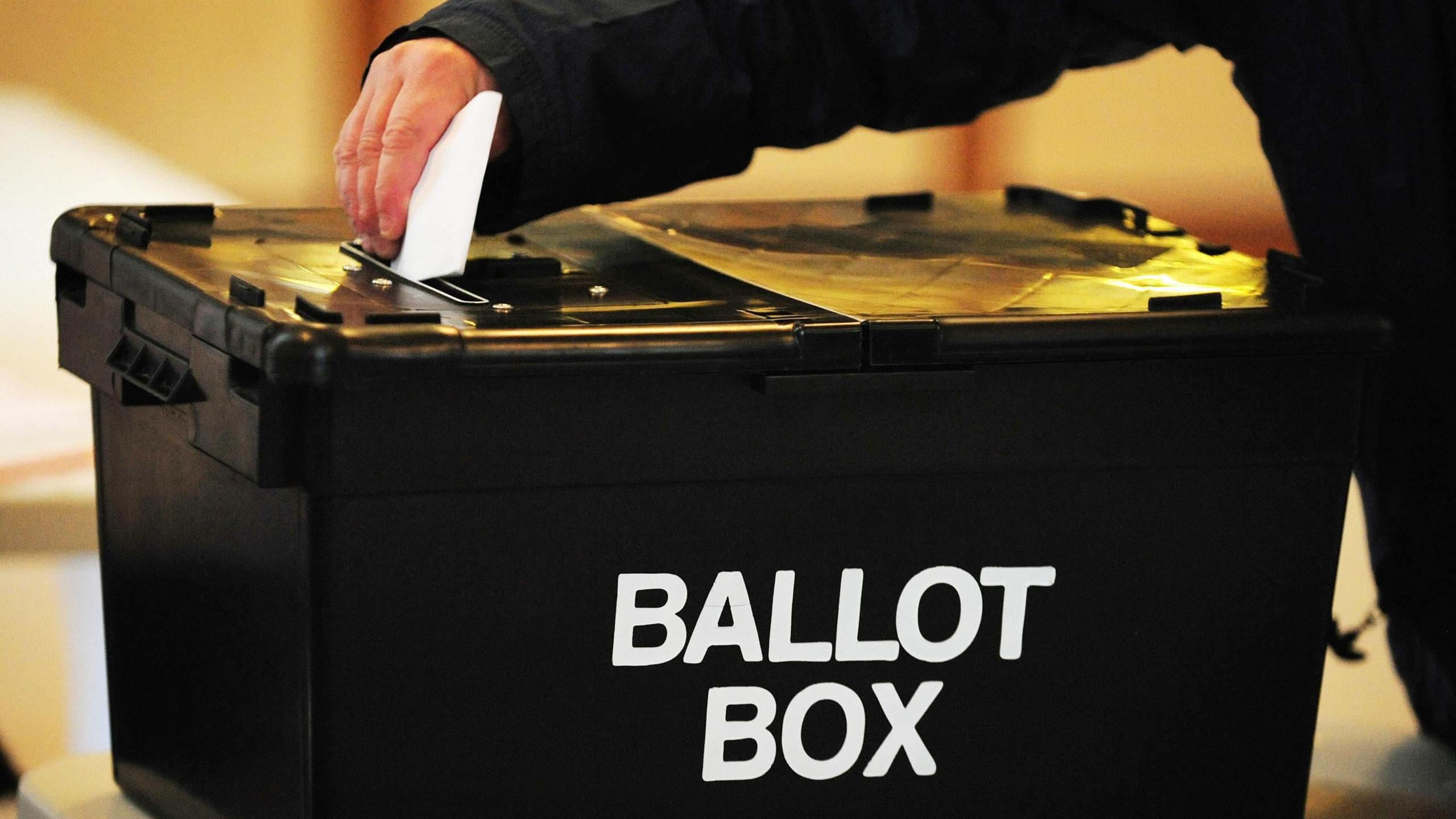 A person wearing a black jacket putting a folded ballot card into a black bin which says 'Ballot Box' on the side. 