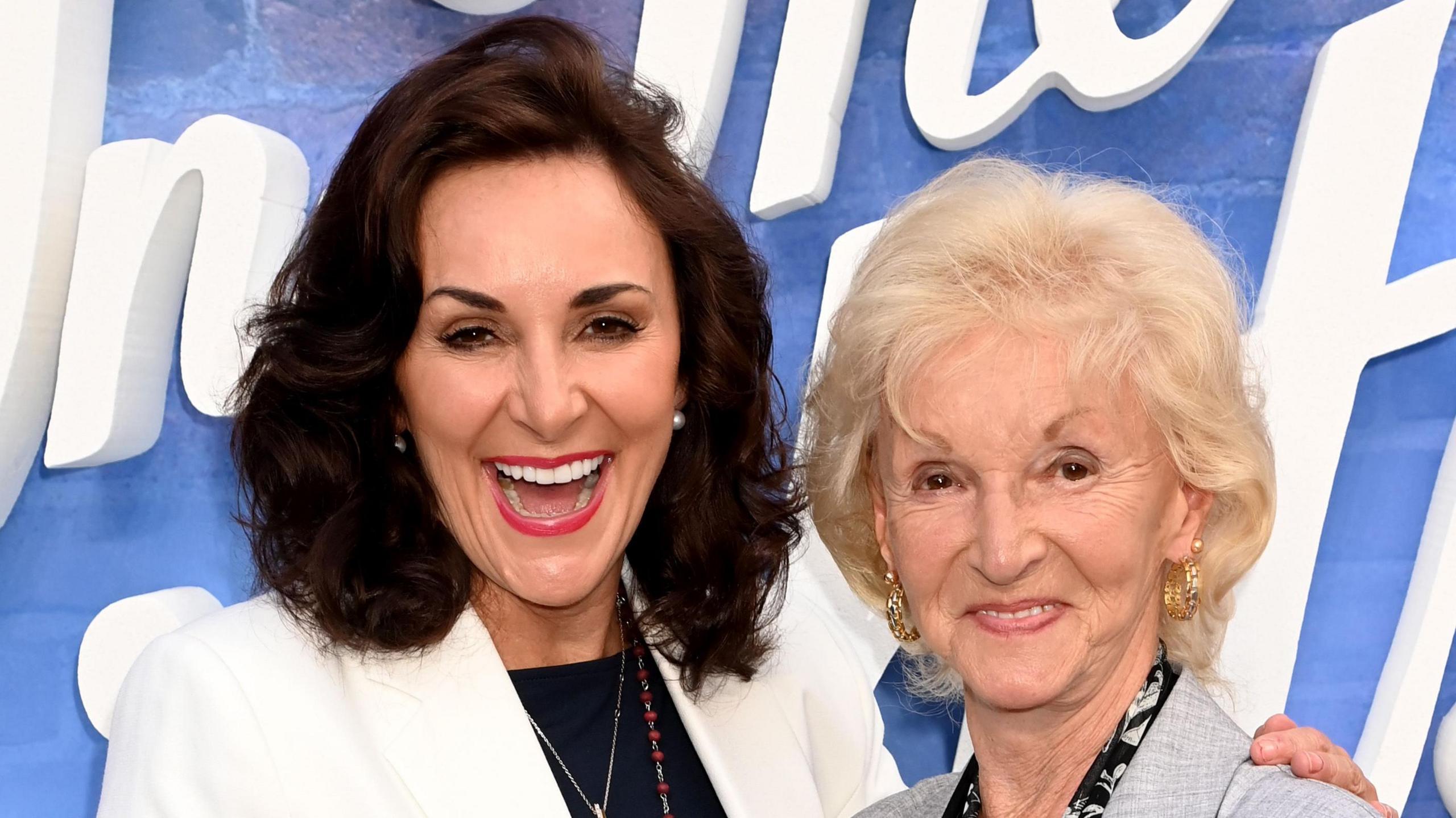 Shirley Ballas and her mother smile as they pose together at a photo call at an event. They stand in front of a blue and white backdrop and both wear smart suit jackets.