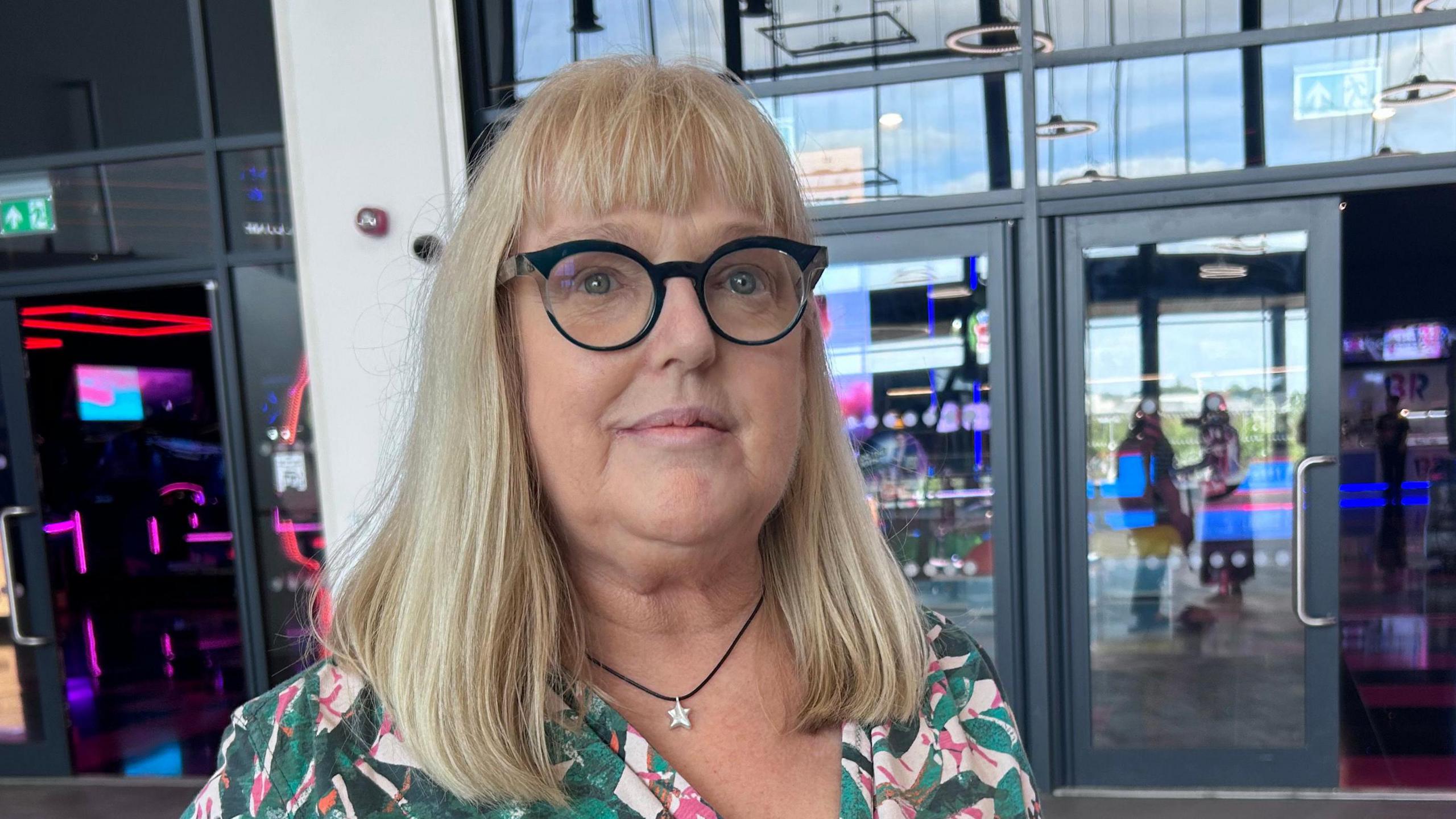 A woman with shoulder length blonde hair and glasses poses at the entrance of a cinema. She has a floral green, pink and white top.