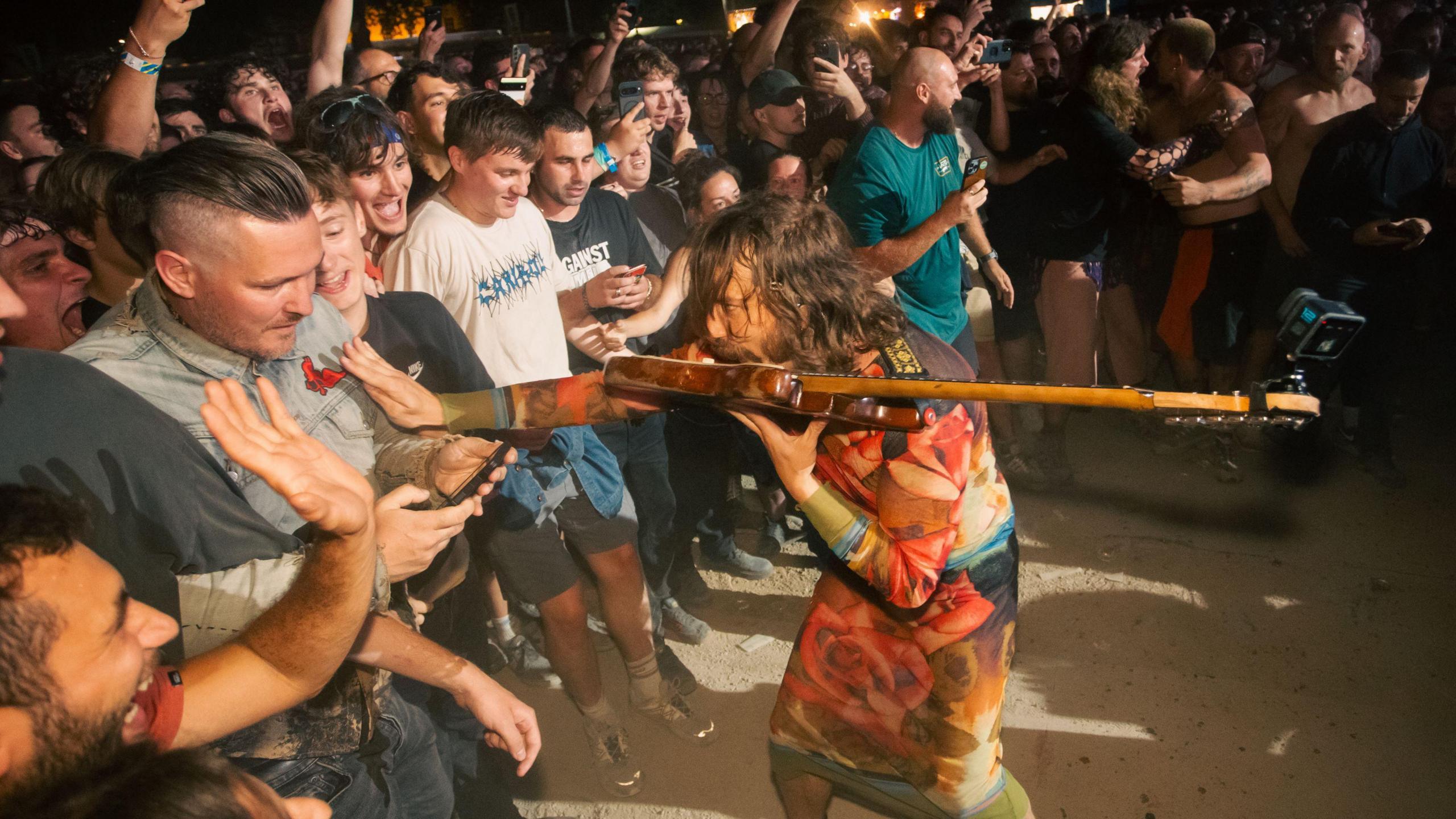 A member of the band Idles plays an electric guitar with his teeth as he stands close to the front row of the audience during the band's Queen Square concert in Bristol