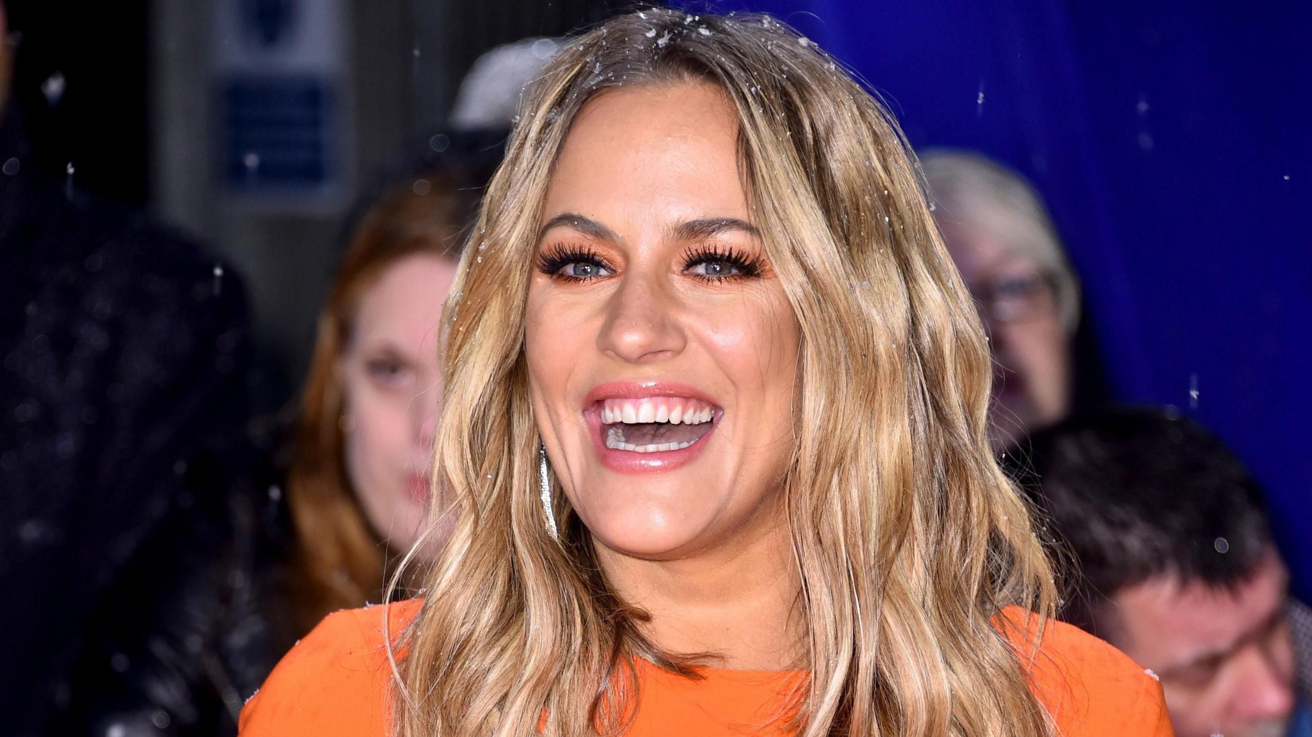 Caroline Flack, a woman who is standing on a red carpet and is wearing a bright orange dress. She has blonde hair and is looking directly at the camera and smiling.
