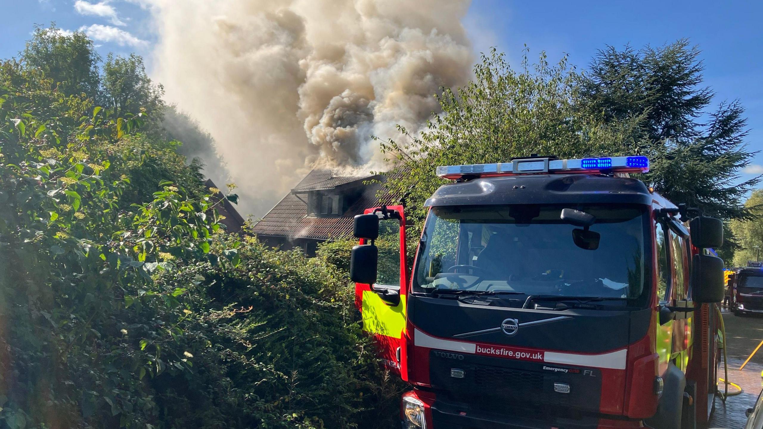 A fire engine is parked on a street; behind it is a line of trees and a house that has huge brown plumes of smoke rising from it into the blue sky.