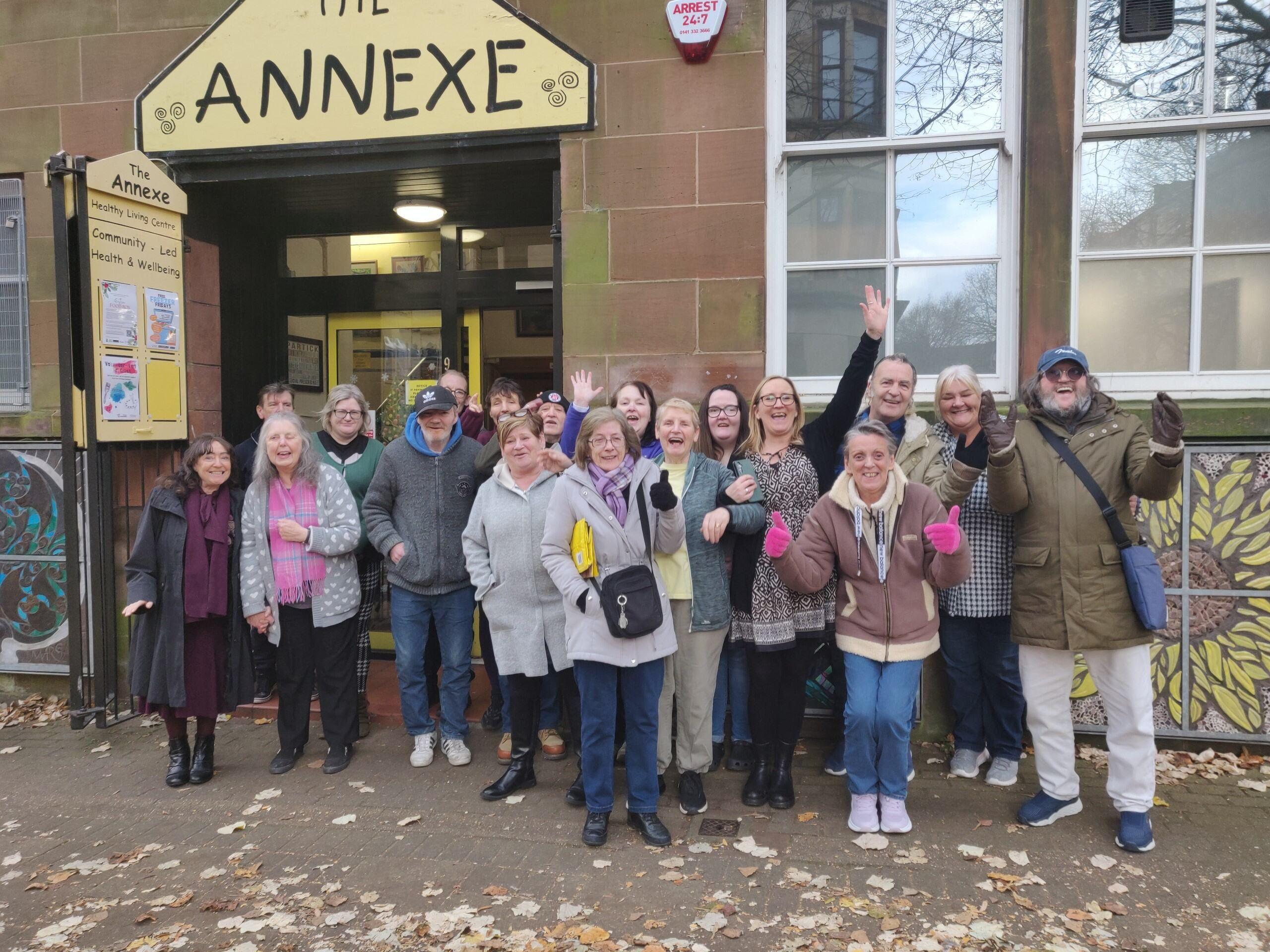 A group of people stand waving and cheering outside a sandstone building with the nameplate The Annexe on it