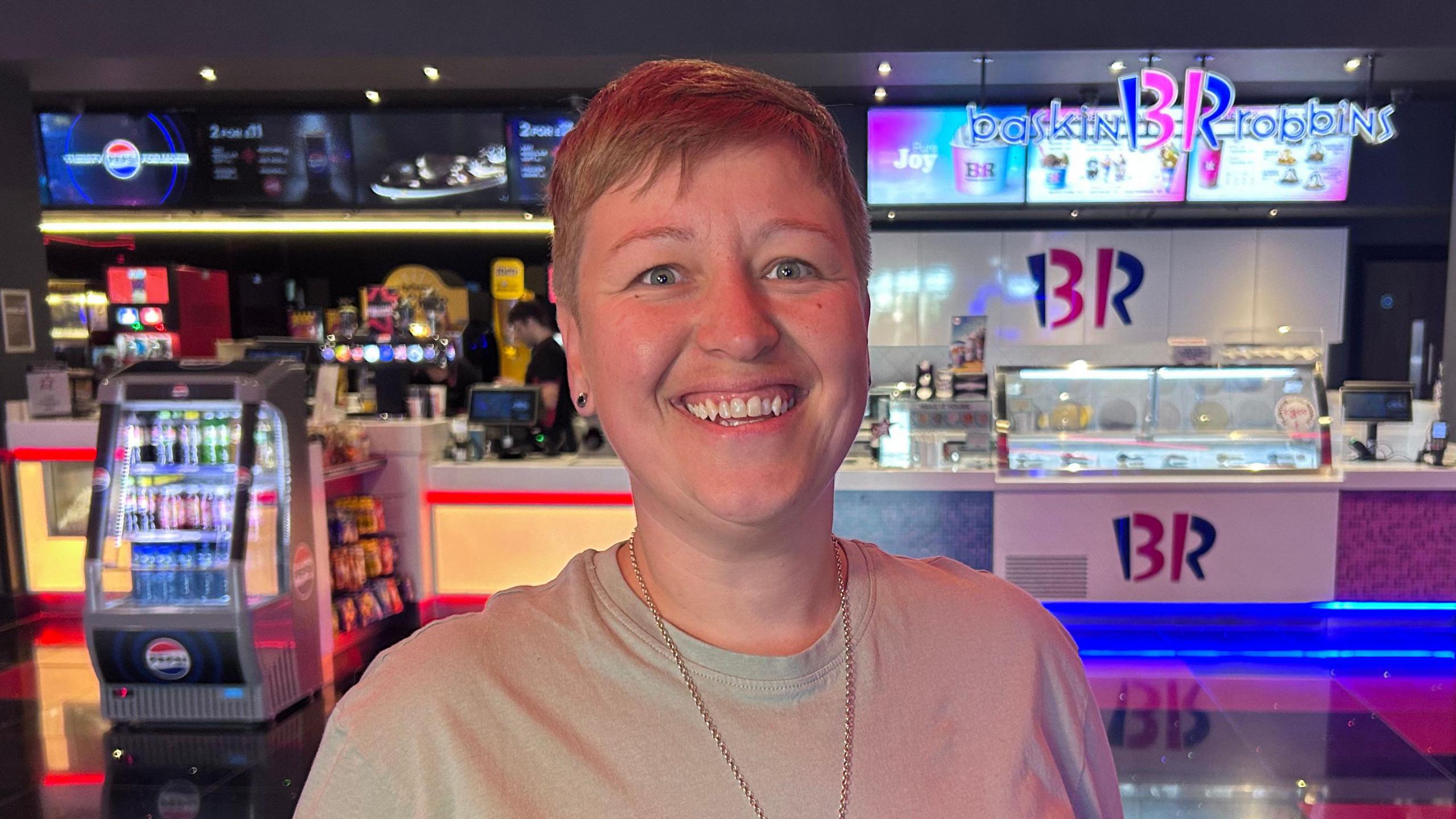 A woman with short fair hair stands in the lobby of a cinema, with refreshments like popcorn and ice cream being sold in the background.