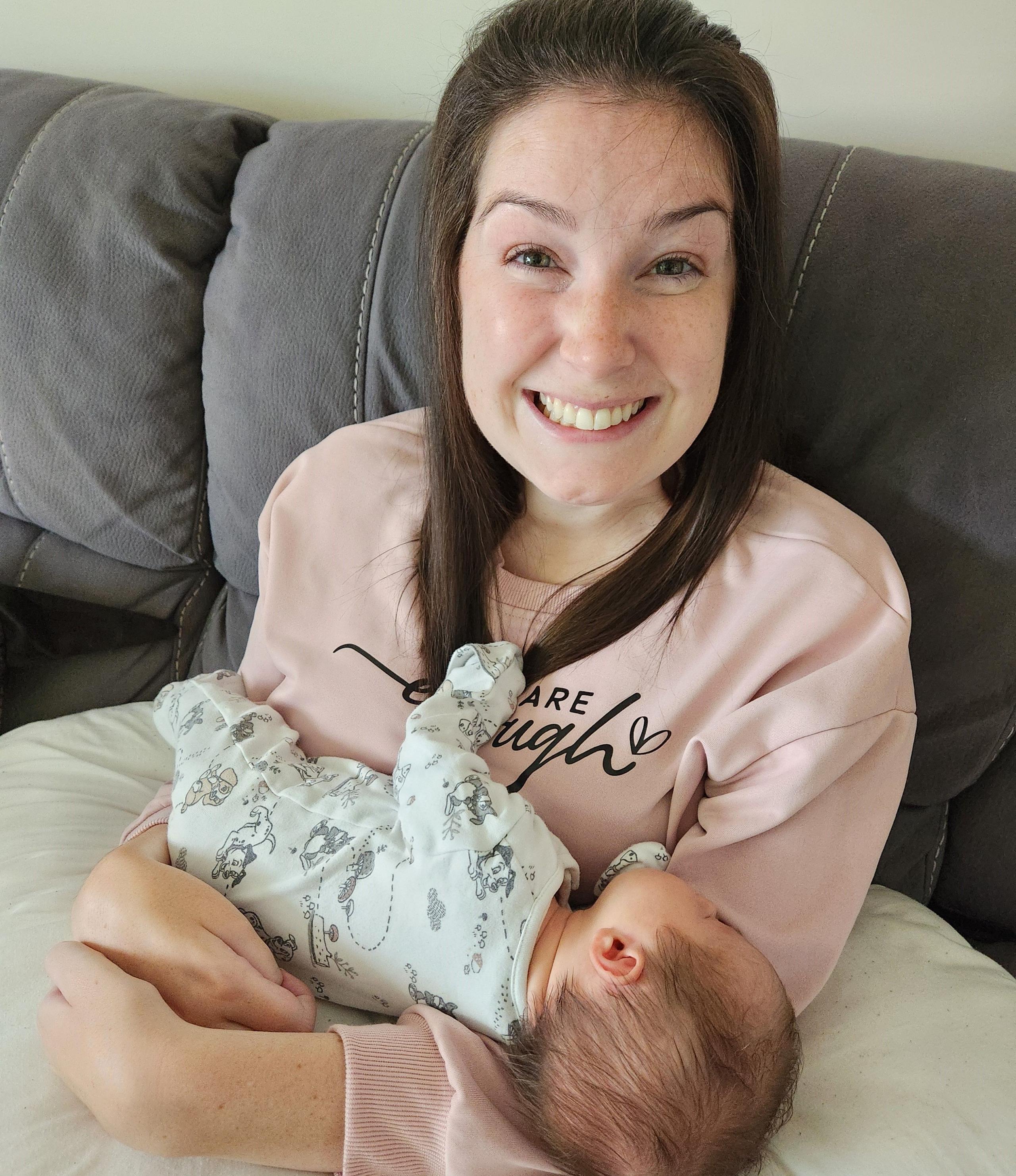 Sitting on a grey sofa and wearing a pink jumper, Lucy Lintott Smith beams as she hold her third baby, who is supported on a maternity cushion. 