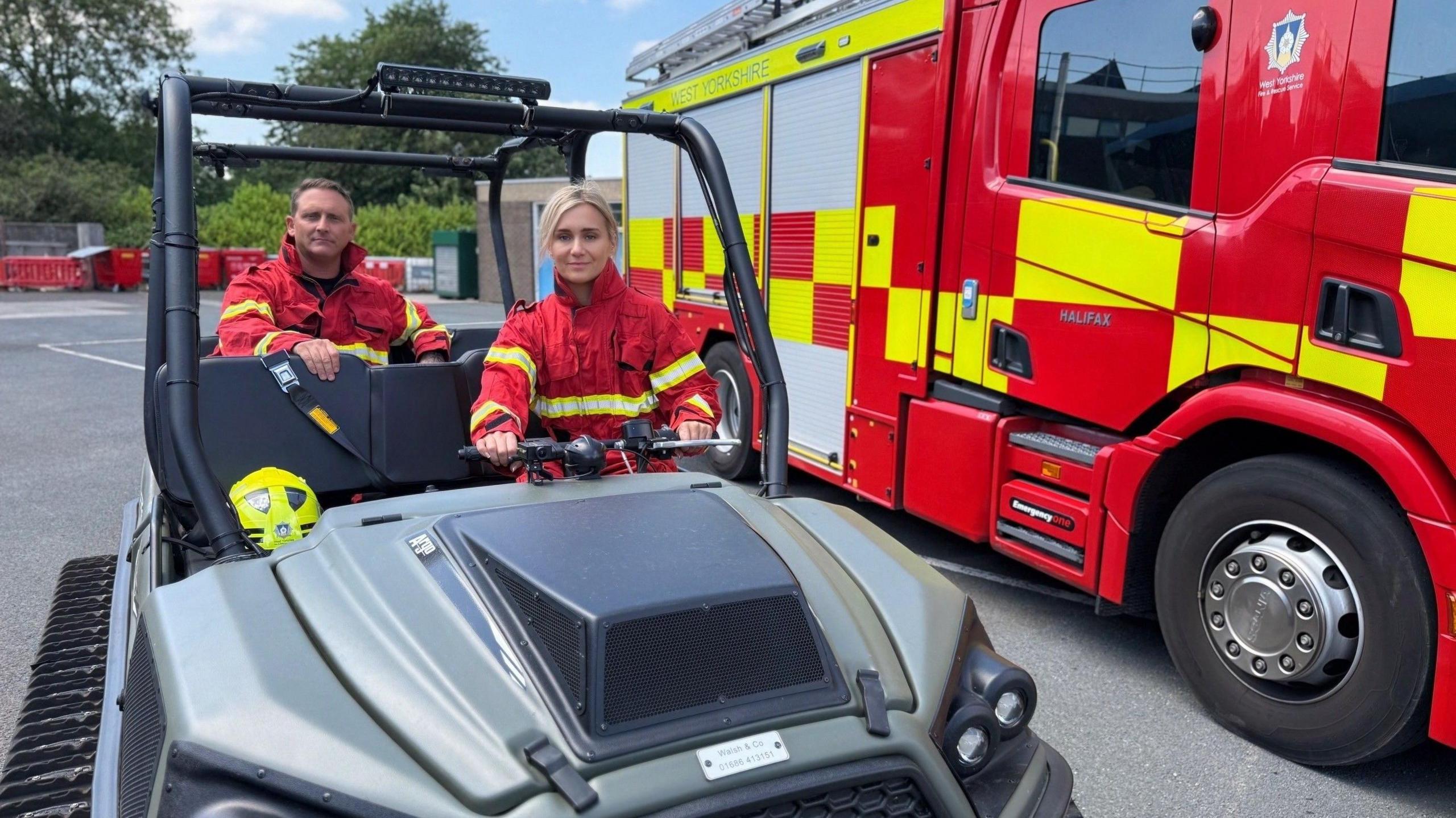Two people wearing red high-visibility rescue jackets sit in a rugged off-road utility vehicle parked beside a red West Yorkshire Fire & Rescue Service fire engine in an emergency services depot. A yellow rescue helmet is visible inside the vehicle, with trees and equipment in the background.