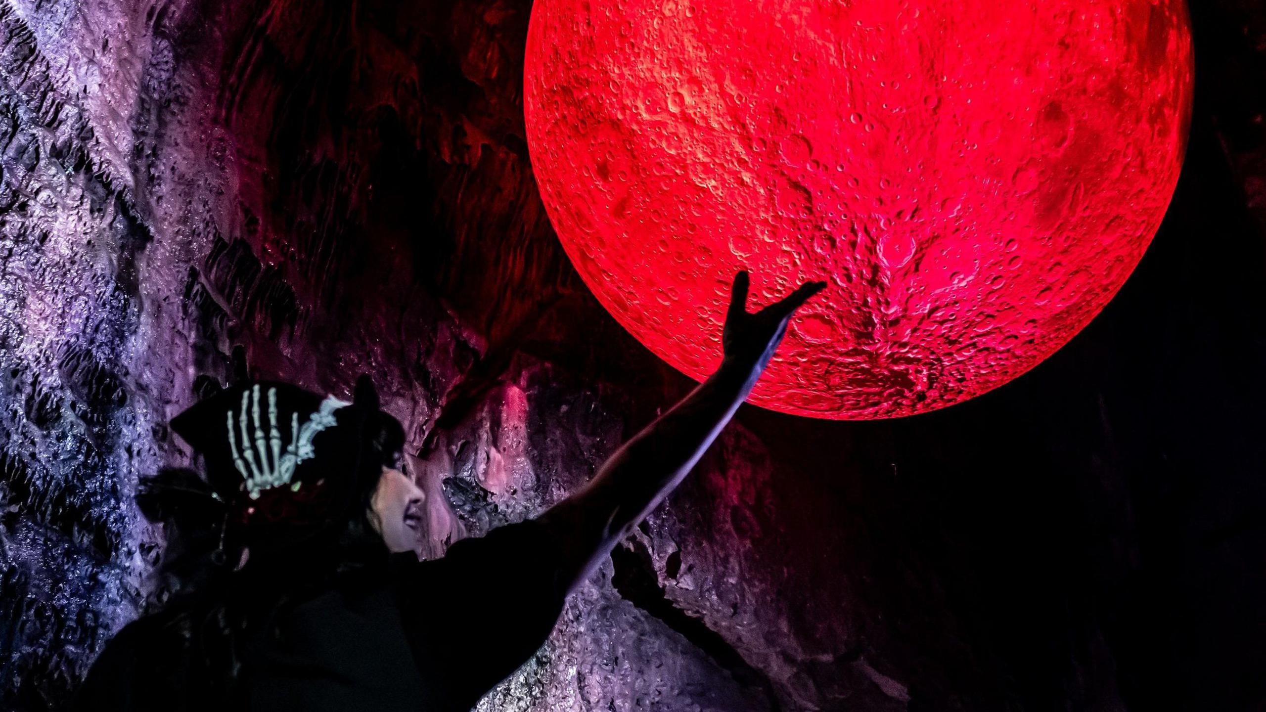 A woman dressed as a witch is silhouetted against a red blood moon artwork inside Wookey Hole caves in Somerset