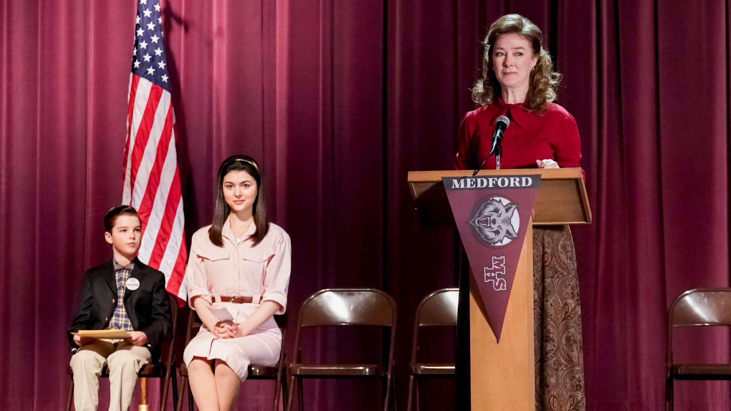 Mahaffey stands at a podium on the set of young sheldon. she is wearing a red sweater and brown skirt. to the side, a young girl and young boy sit on folding chairs. there is a purple curtain behind them and an american flag.