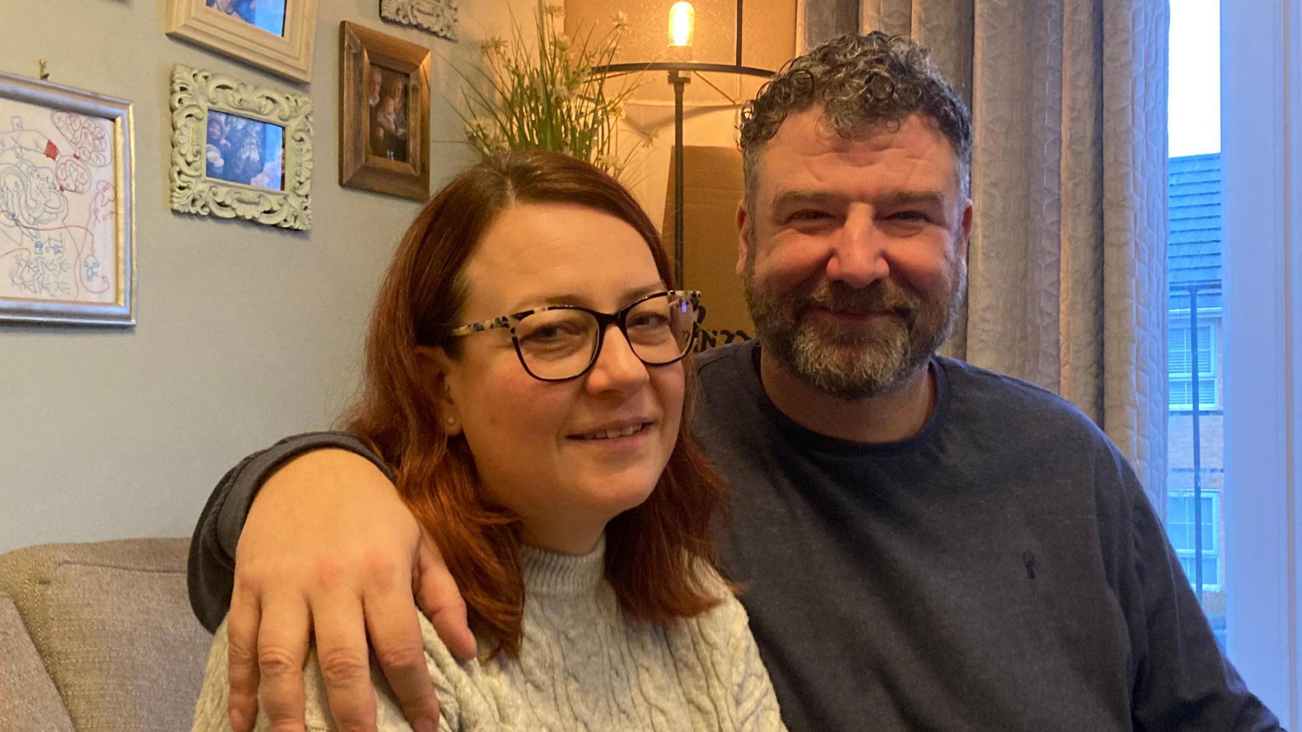 Kirsty and Donovan Shears sitting on a sofa, together at home. There are picture on the wall and they are both wearing jumpers and he has his arm around her shoulders.