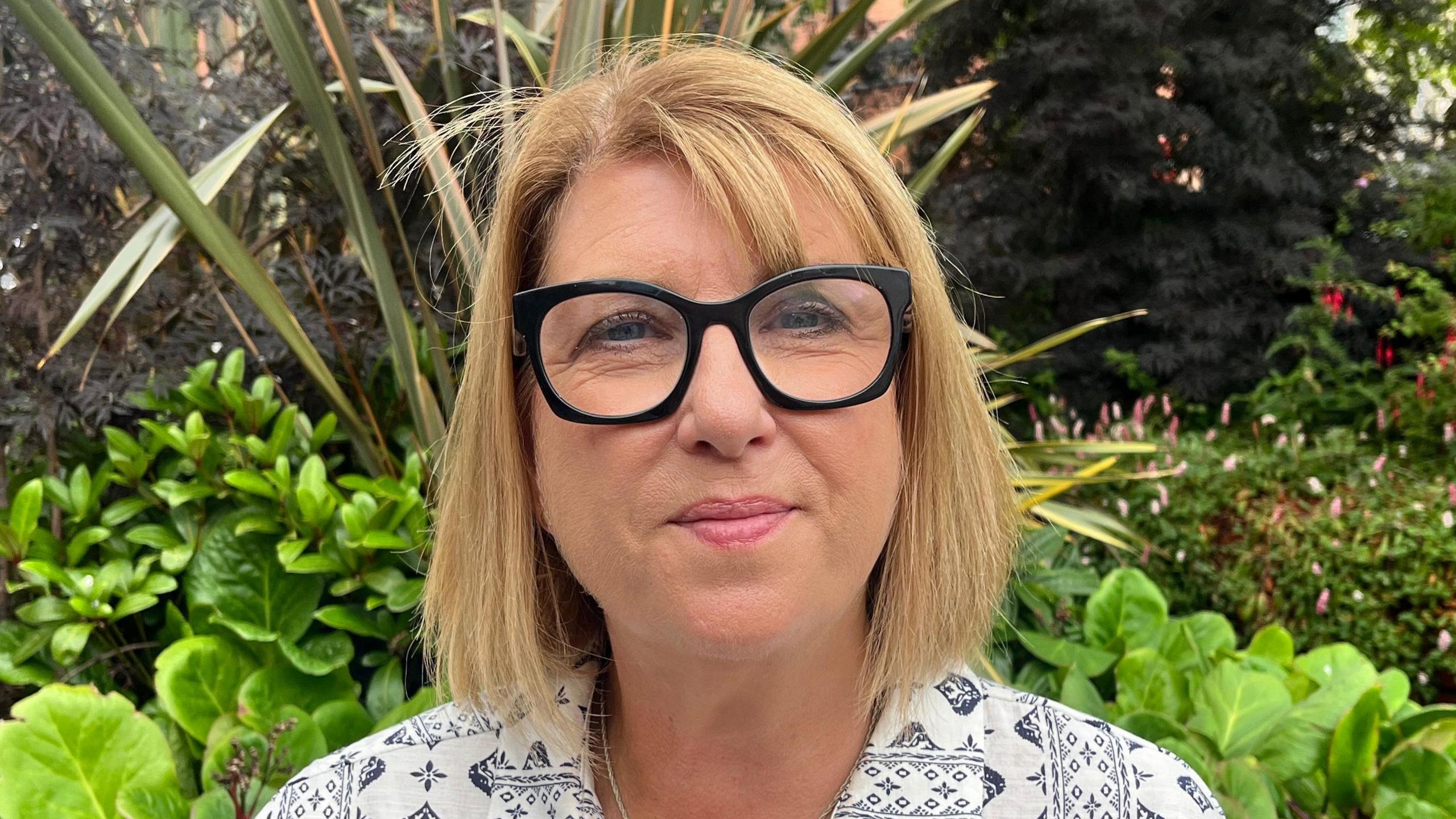 A woman with chin-length blonde hair stares at the camera as she wears black glasses and pink lipstick. She is wearing a white shirt with a black pattern on it. Behind her is flowerbed with green plants in it.