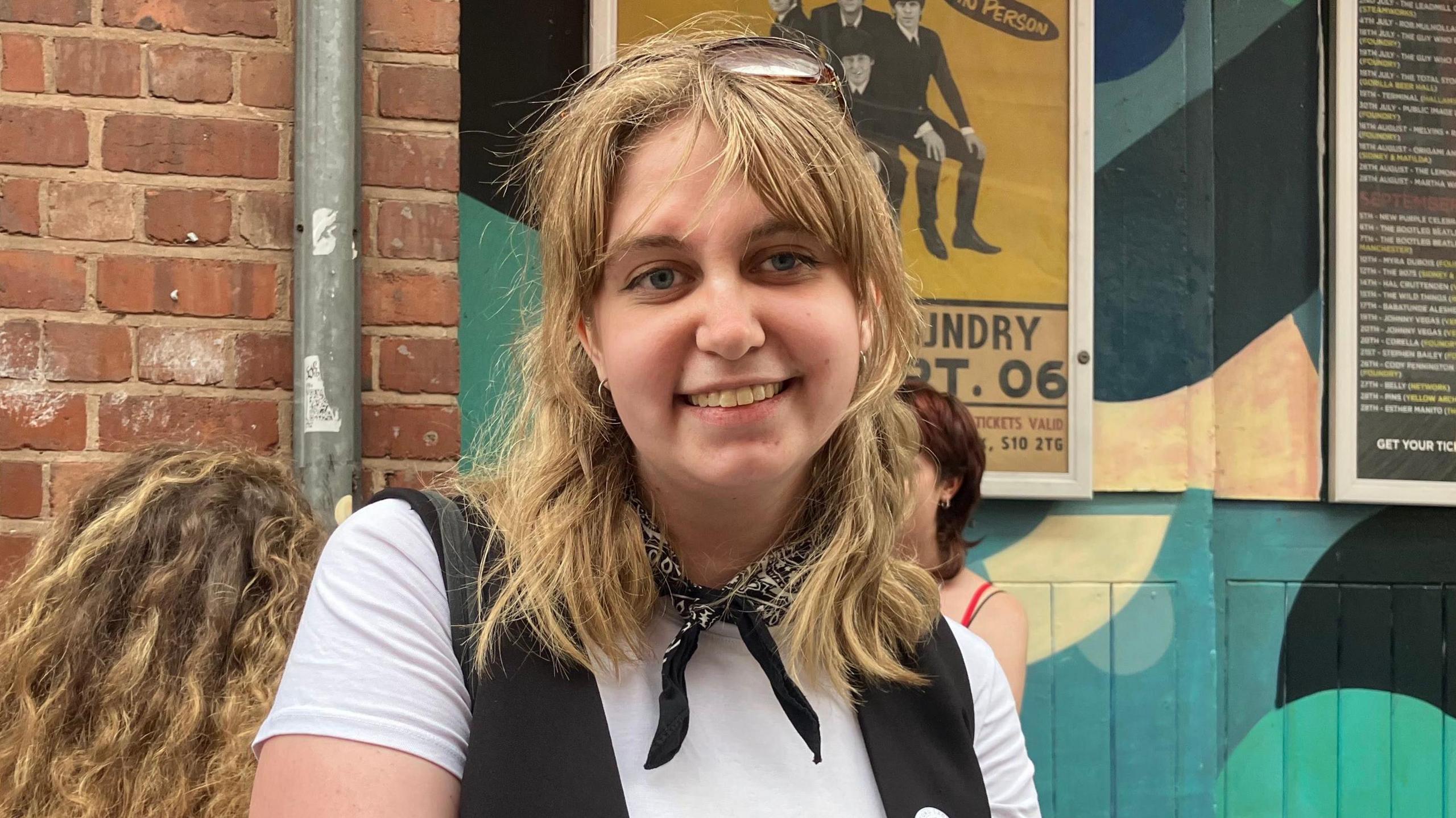 A woman with blond shoulder length hair. She wears a white t-shirt, black cotton tux, and black bandana around her neck.
