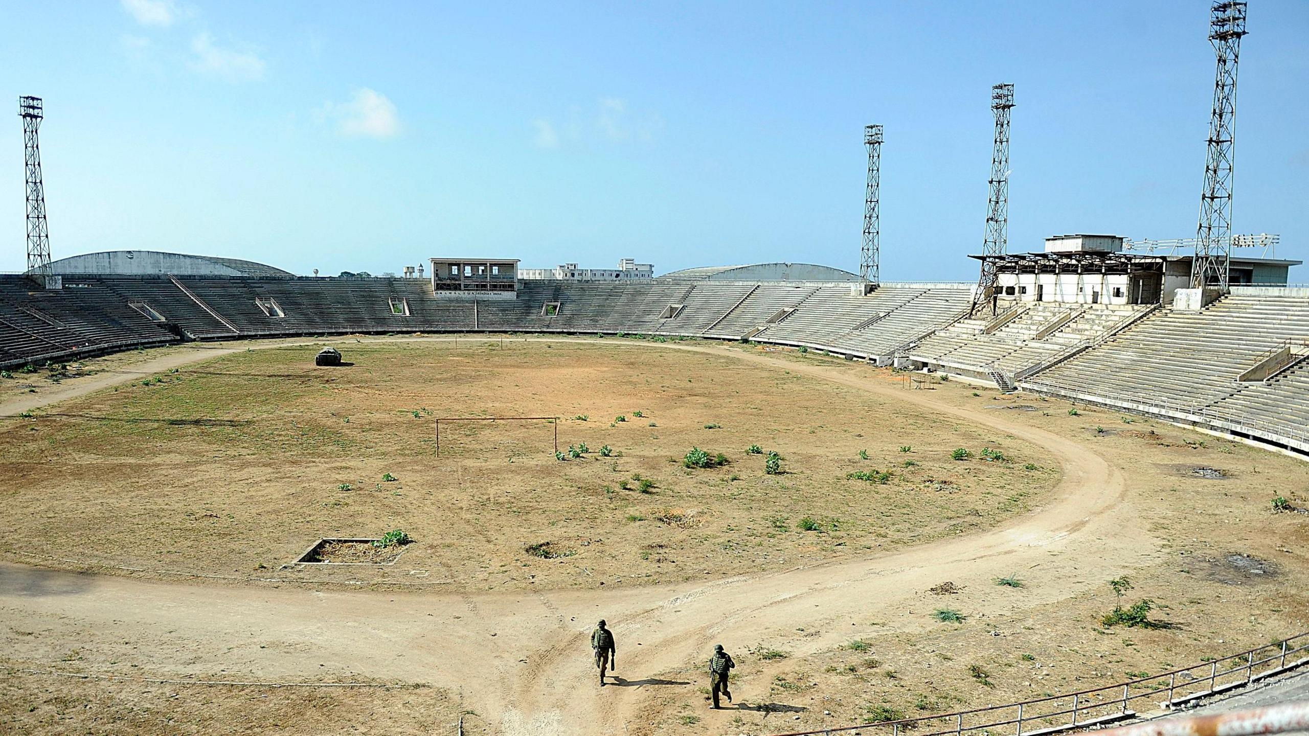 A picture taken from high up in a stand makes two peacekeepers in the foreground appear very small as they stand in a barren and dusty Mogadishu Stadium with low empty stands in the background and floodlight pylons rising into a blue sky