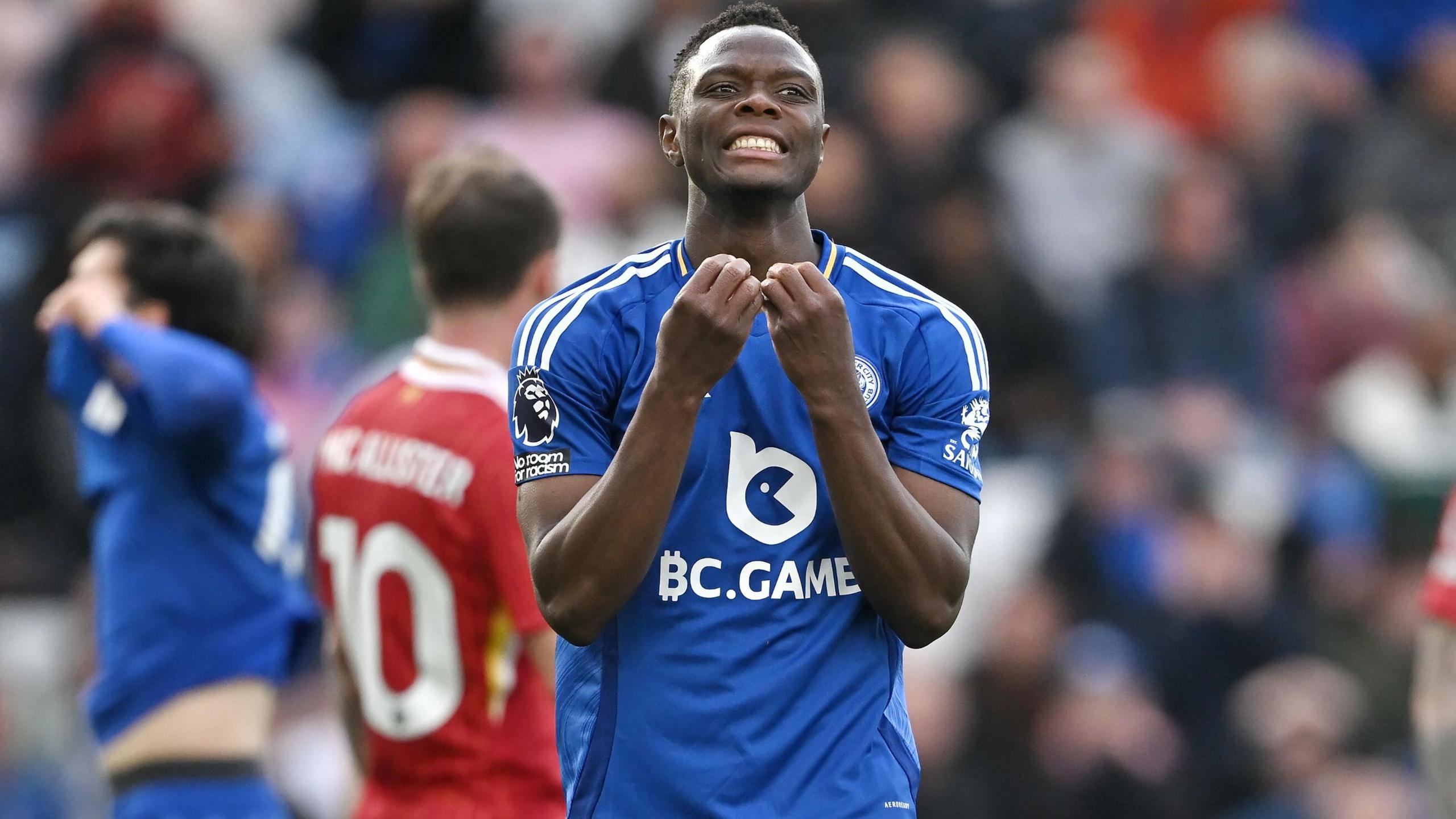 Patson Daka, wearing Leicester's 2024-25 blue home kit, looks on with anguish after the Foxes were relegated from the Premier League following defeat to Liverpool. Blurred Liverpool players wearing red can be seen in the background, as well as a dejected Leicester player with their hands on their face.