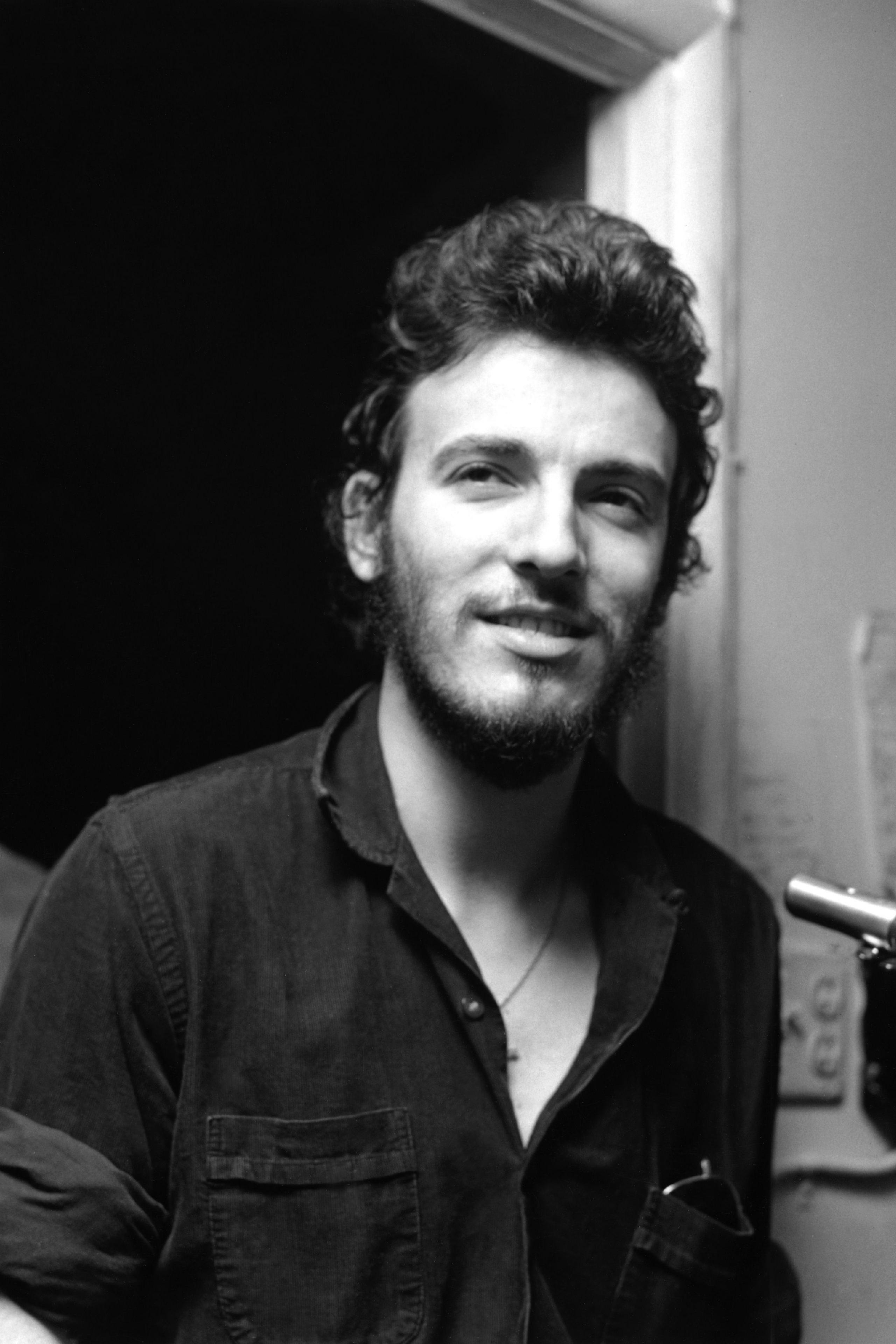 Black and white photograph of Bruce Springsteen backstage at a concert, circa 1970. He has short curly hair and a close-cropped beard and moustache. Springsteen is wearing an open-necked black shirt.