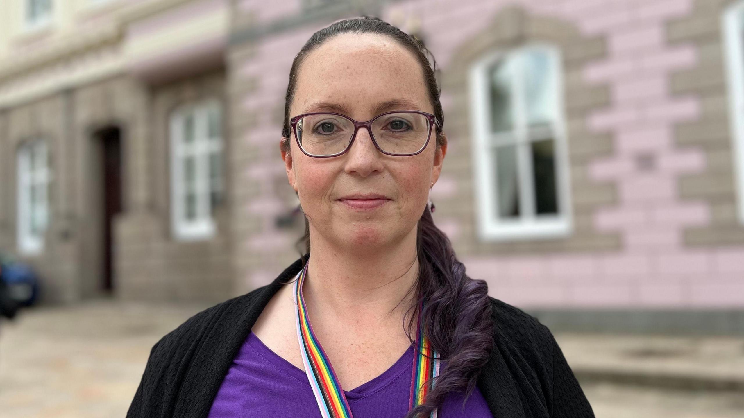 A woman in a purple top and glasses stands in front of a pink granite building.