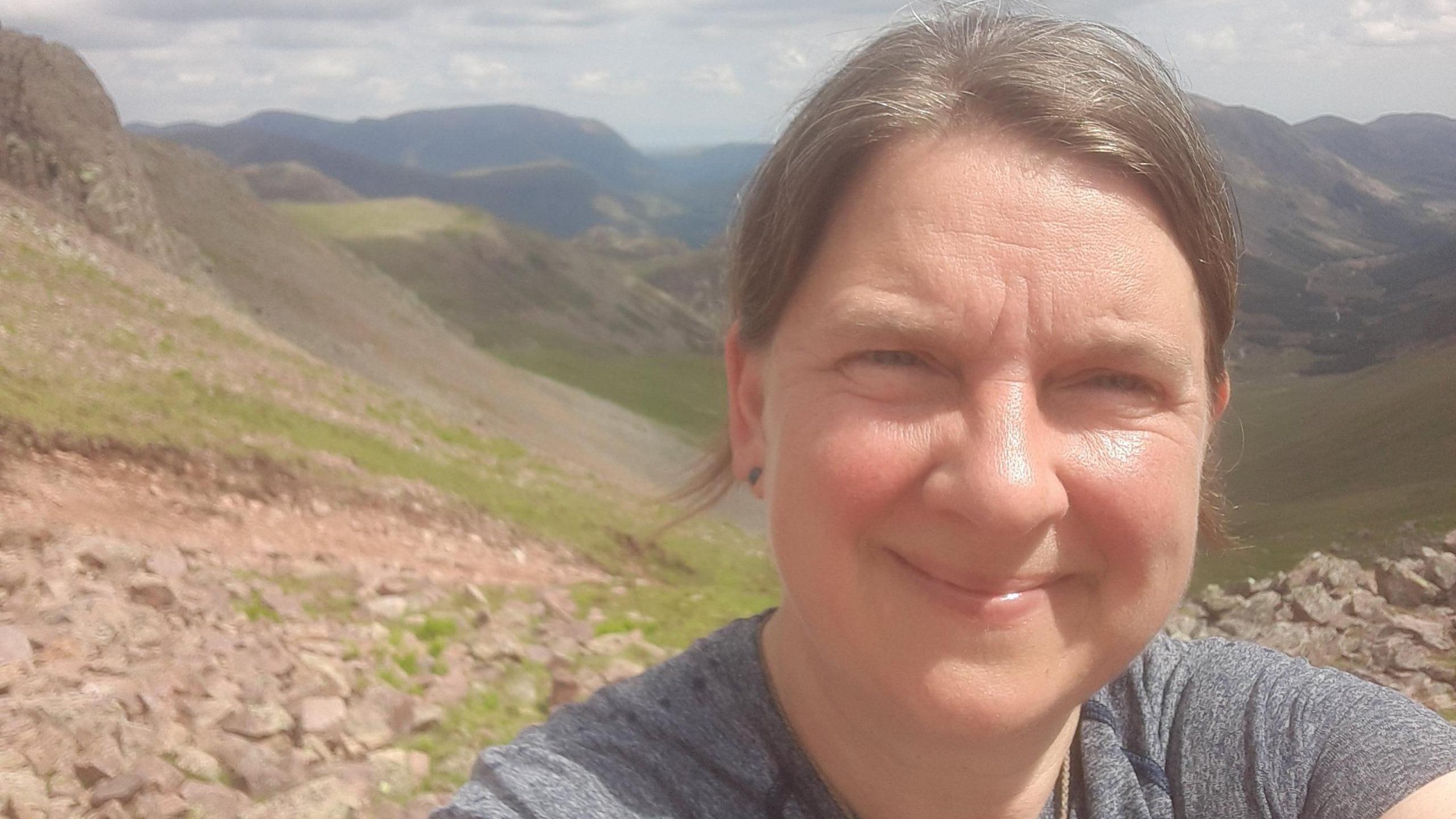 A close-up picture of Zoe Gilbert. It is a selfie taken in the fells. She has her light brown hair tied back and is smiling. She has blue eyes. Behind her is the side of a fell with patches of grass and rocks. Further in the background is a valley and other fells.