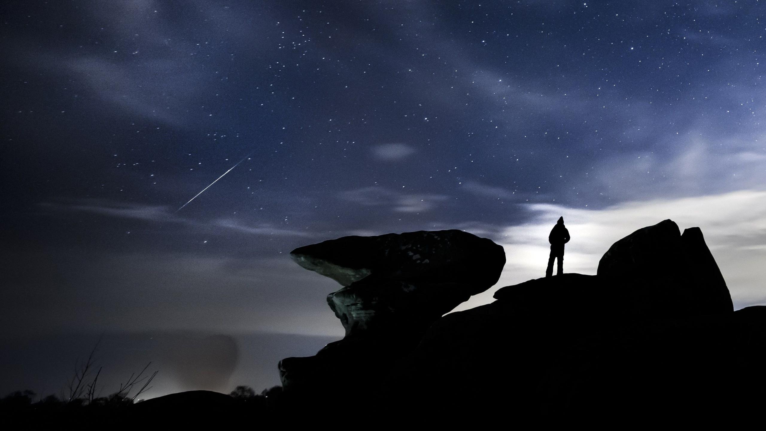 A silhouetted figure stands on rocks and watches a meteor streak across the sky 