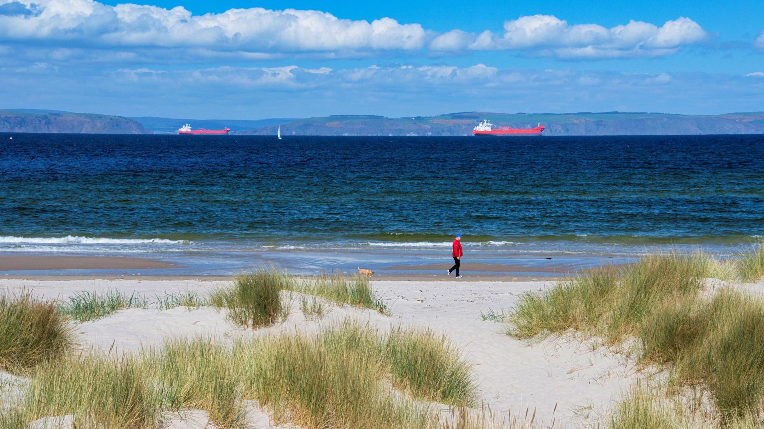 A person wearing a red jacket, black trousers and white trainers walks along Nairn beach. Out at sea are two red ships.