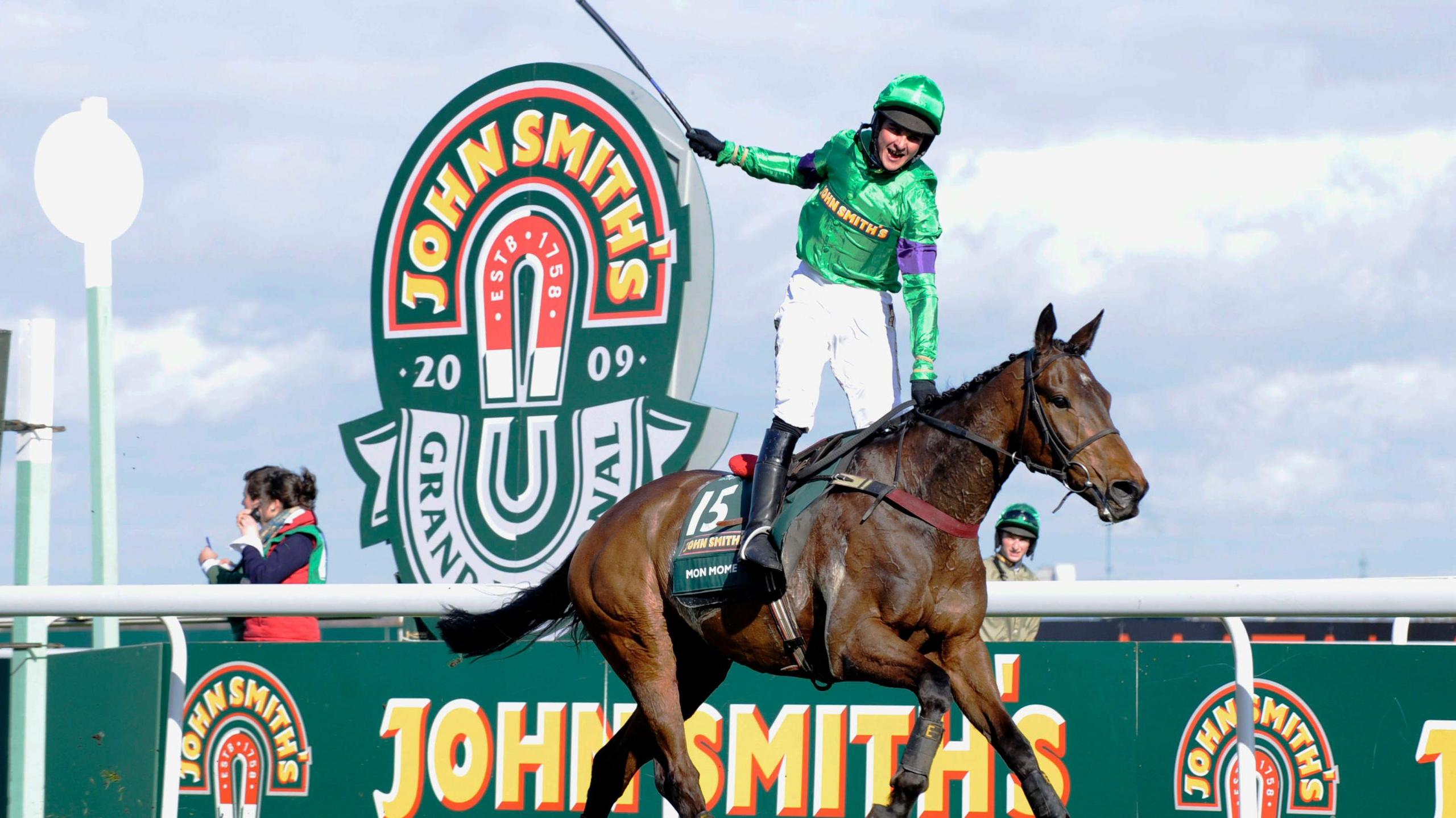 A jockey wearing a bright green jersey and helmet with white trousers and black boots rides a brown horse. 