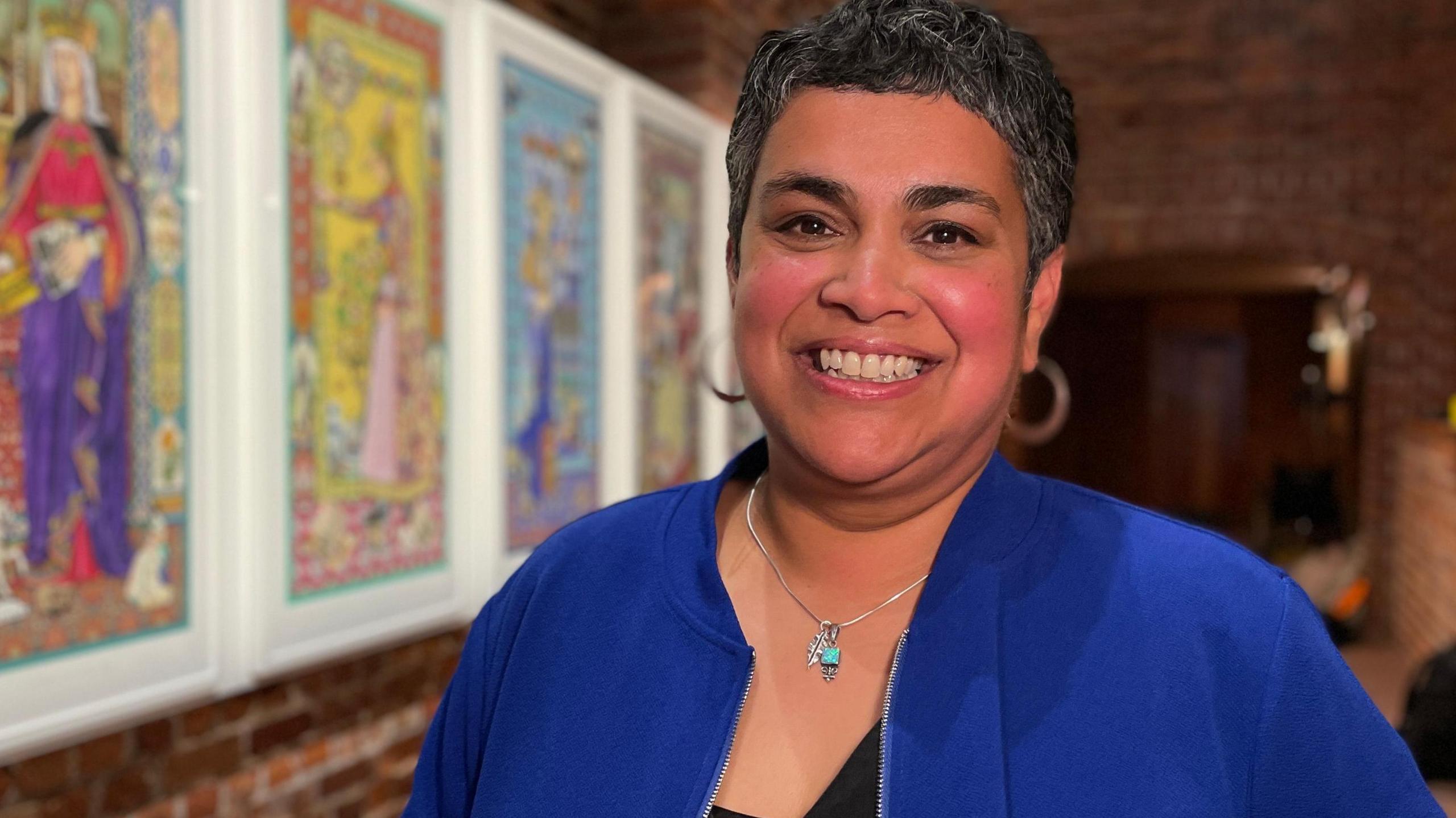 Devi Singh is smiling at the camera. She has short cropped hair and is wearing a cobalt blue bomber-style jacket over a black top. She is also wearing hooped earrings and a silver necklace with a turquoise pendant.
