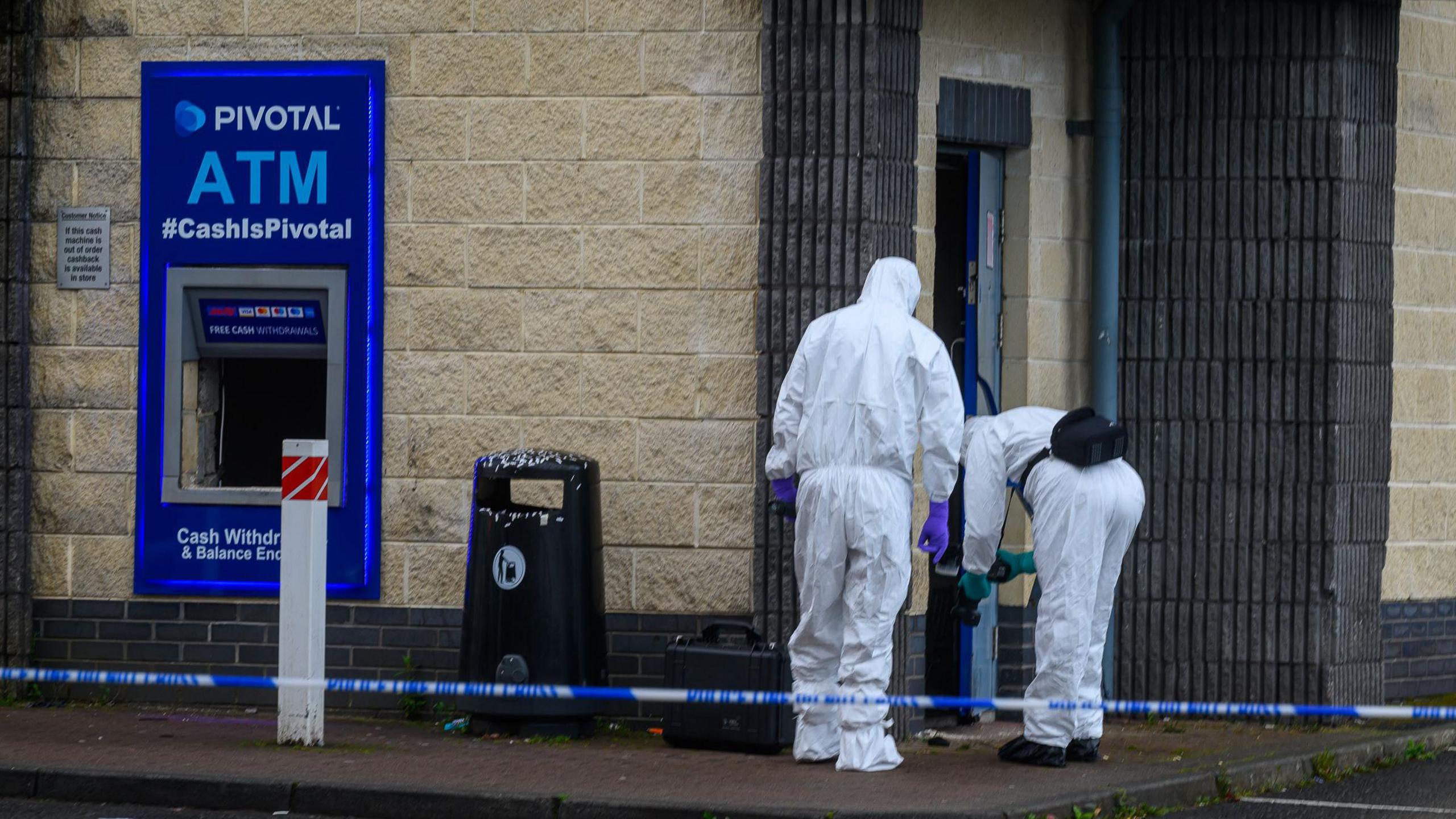 ATM missing from wall of shop with men in white suits investigating.