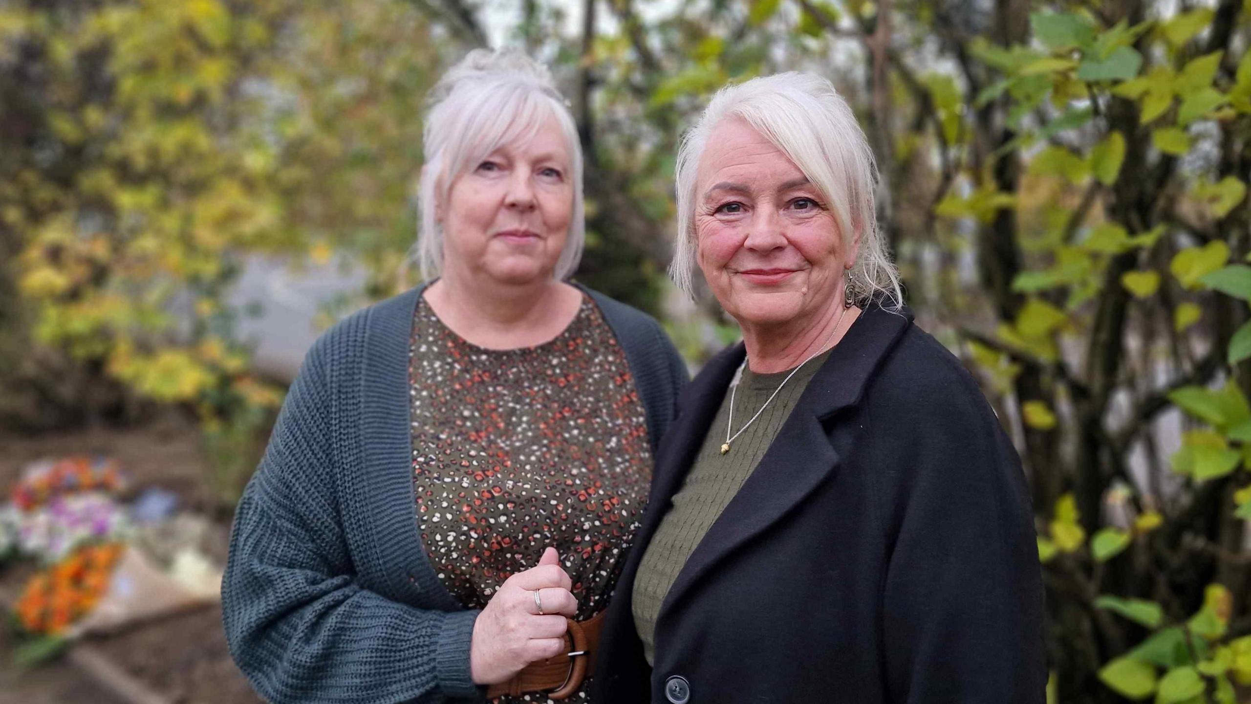 Two women with white hair, one with a cardigan and patterned top, the other with a dark coat and green jumper with a necklace. There are bushes in the background.