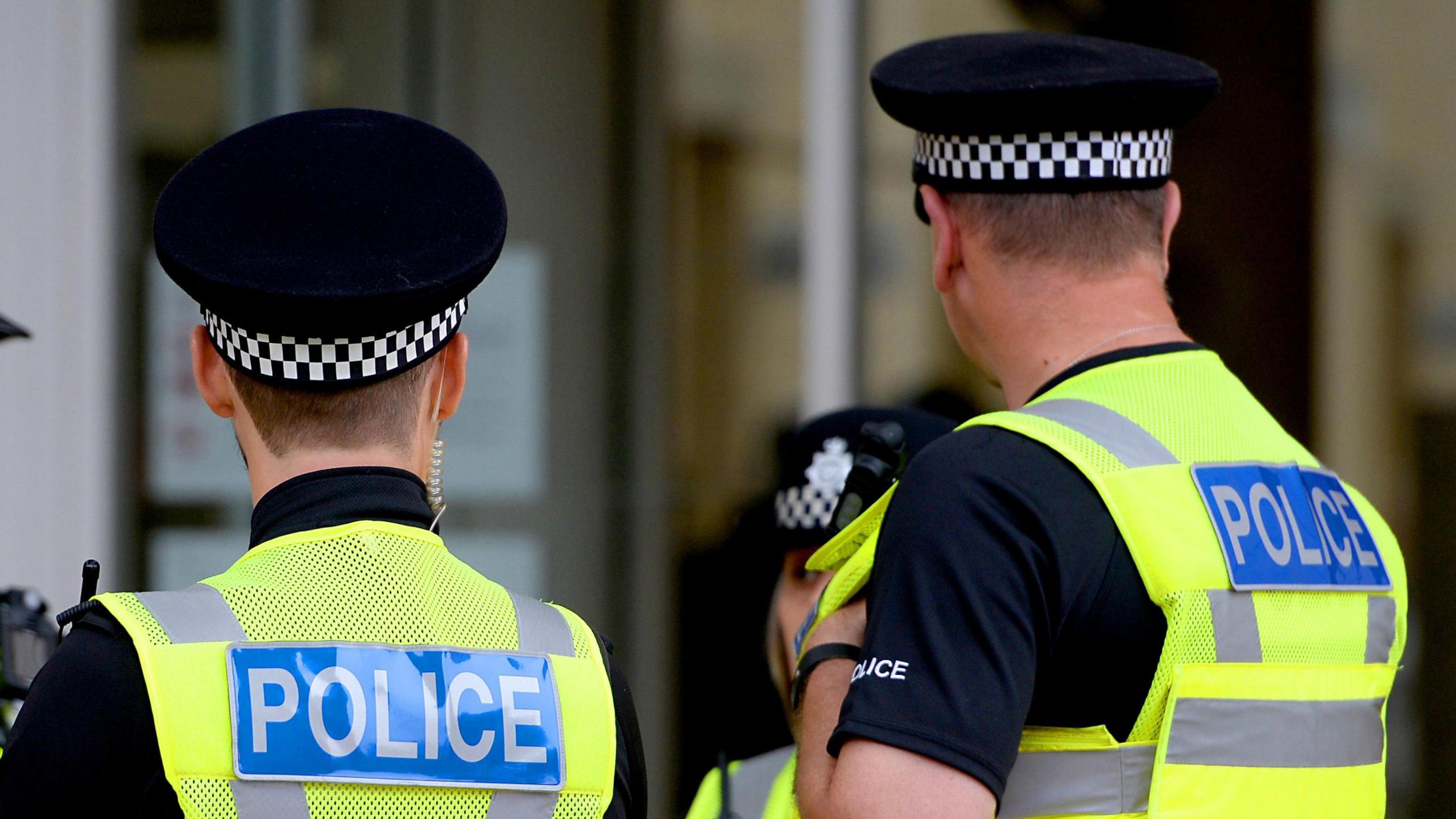 Two police officers face with their backs to the camera, wearing yellow hi-vis jackets with the wording 'police' on the back in blue and white