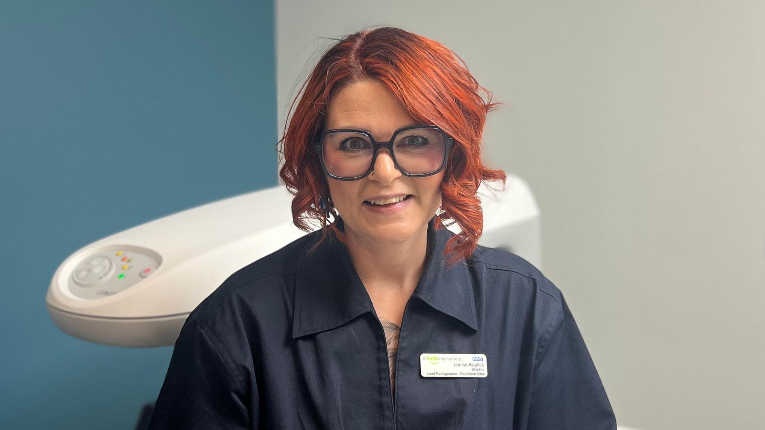 A women with black glasses and short red hair sat down. She is wearing a navy blue shirt. She is in a hospital room with a bone density scanner behind her.