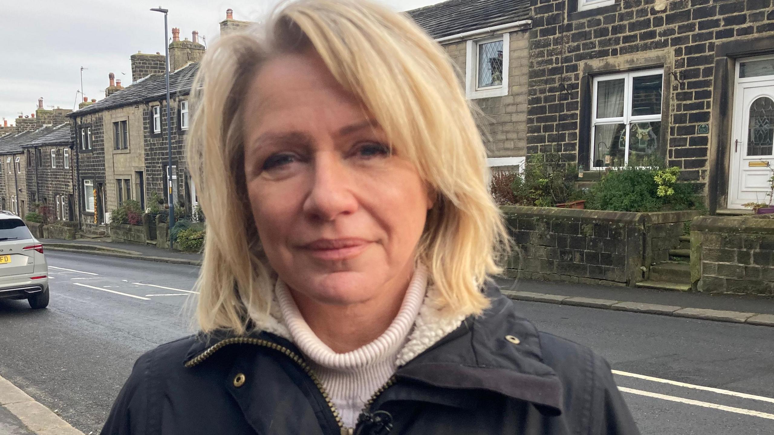 A woman in a dark jacket and with blond hair looks at the camera